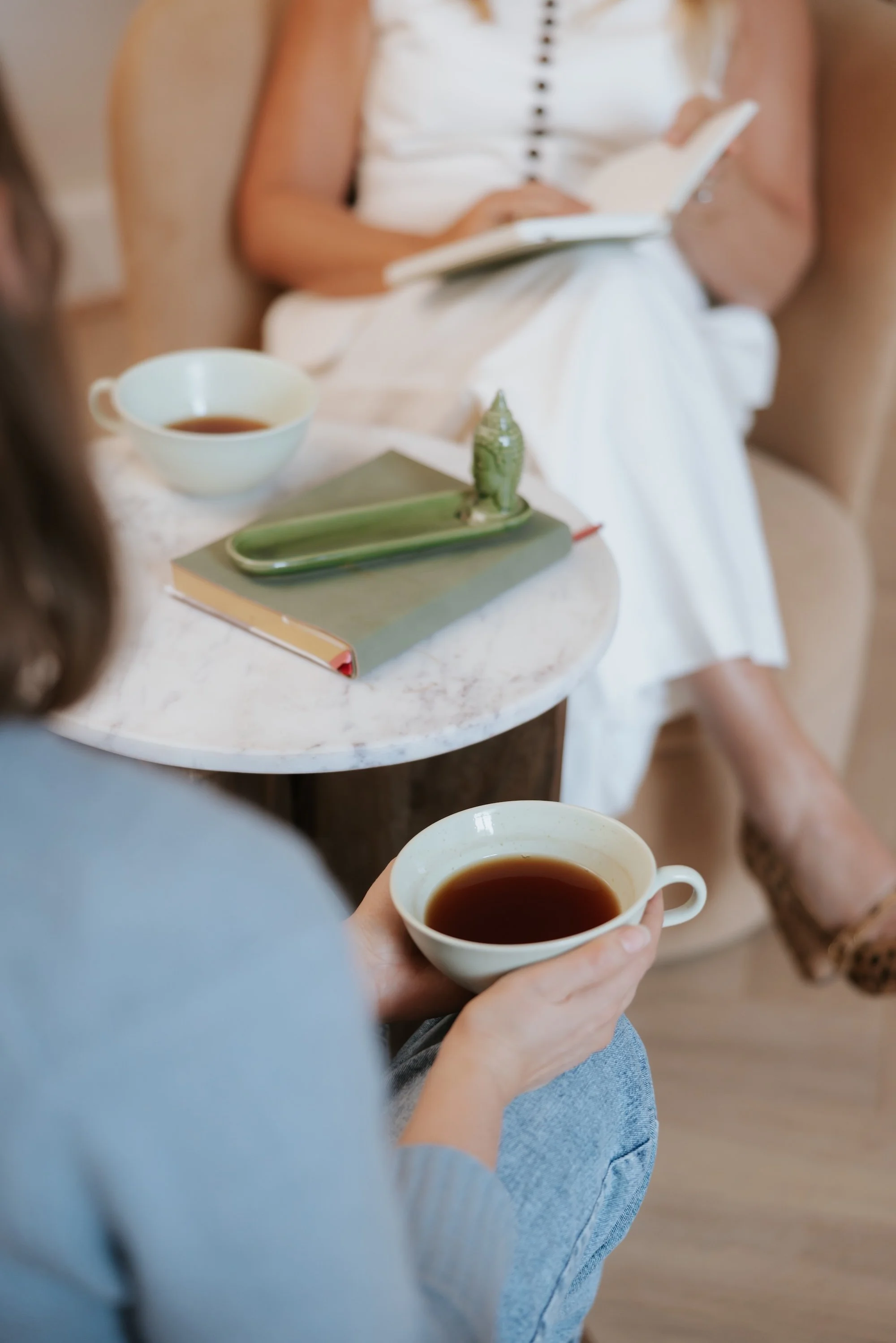 A person holding a cup of coffee in their hand, with a table and another cup of coffee in the foreground. In the background, a person wearing a white dress is reading a book, sitting on a beige chair.