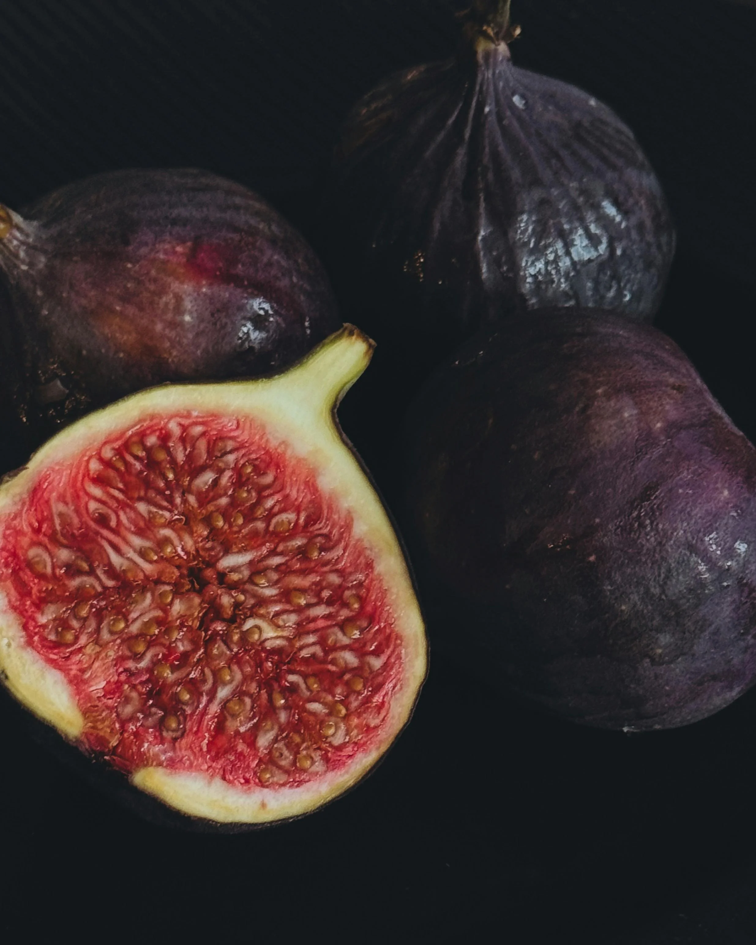 Close-up of a halved and whole purple fig with red interior on a black surface.