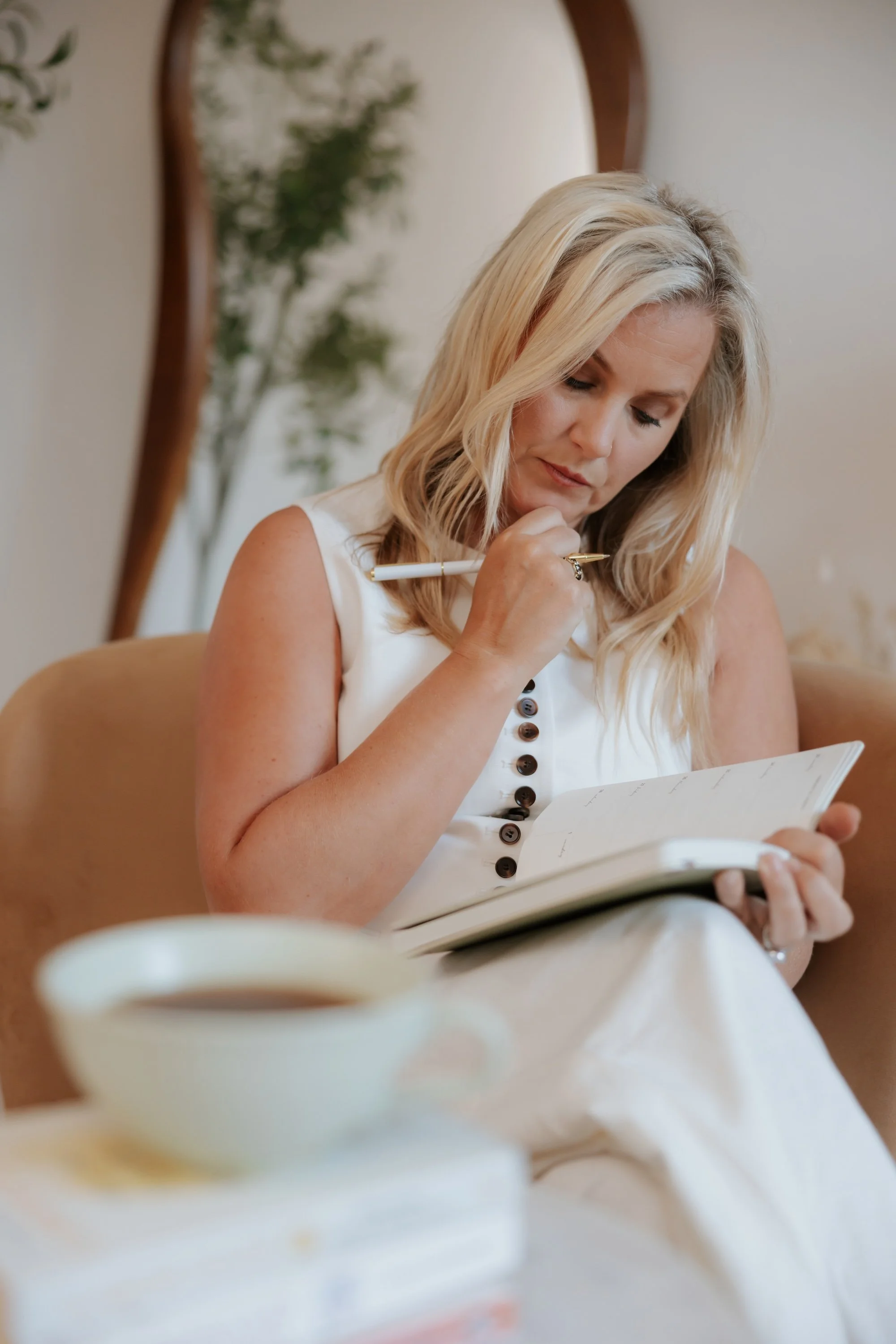 A blonde woman in a white sleeveless top is sitting indoors, holding a pen in her right hand and a notebook in her left, appearing to write or draw while looking down.
