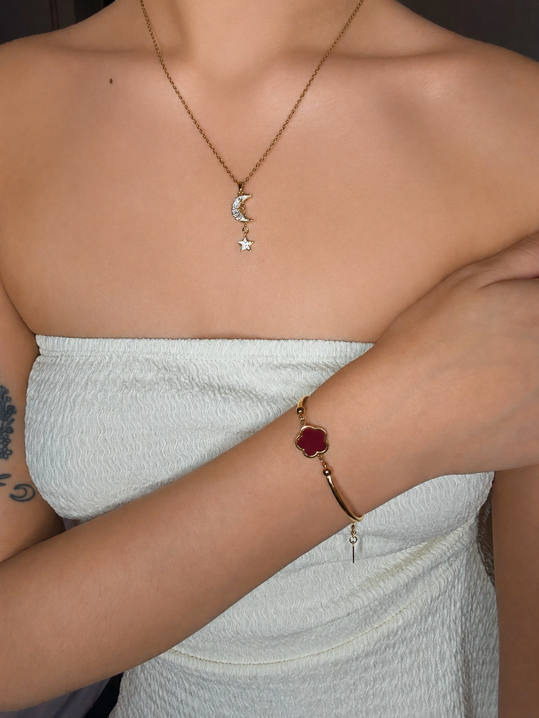 A woman wearing a gold necklace with a crescent moon and star pendant, and a gold bracelet with a red four-leaf clover charm on her wrist.