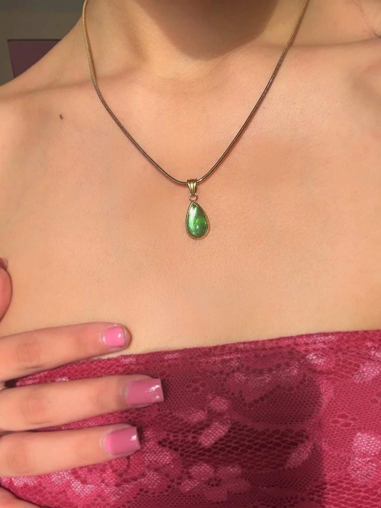 A woman wearing a necklace with a teardrop-shaped green gemstone pendant. She has pink fingernail polish and is dressed in a red lace garment. Her hand is placed on her chest, partially covering her skin and lace clothing.