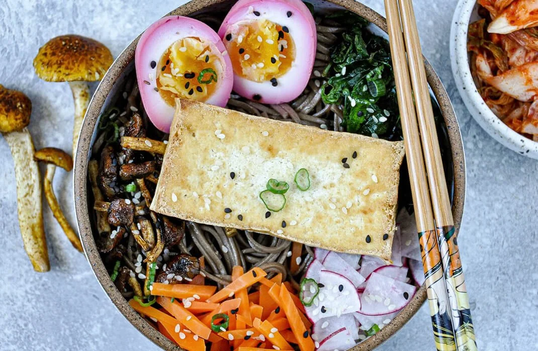 Bowl of ramen with pink egg halves, greens, shredded carrots, radishes, toasted tofu, and chopsticks.