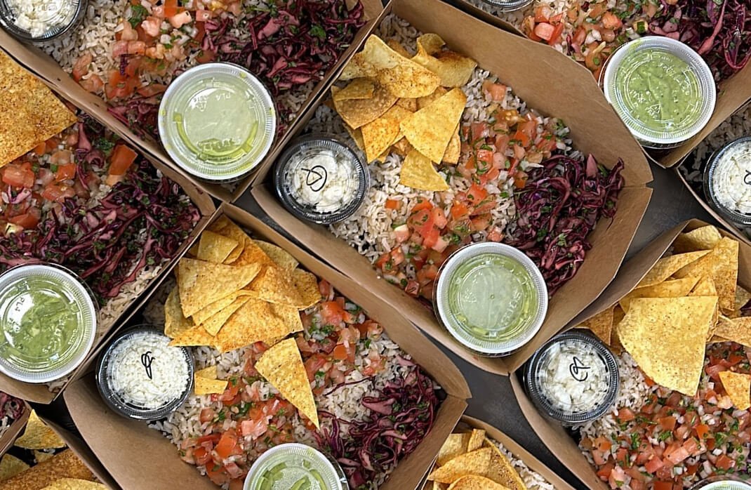 Multiple to-go boxes of Mexican rice, shredded purple cabbage, diced tomatoes, chips, and small containers of green salsa and sour cream.