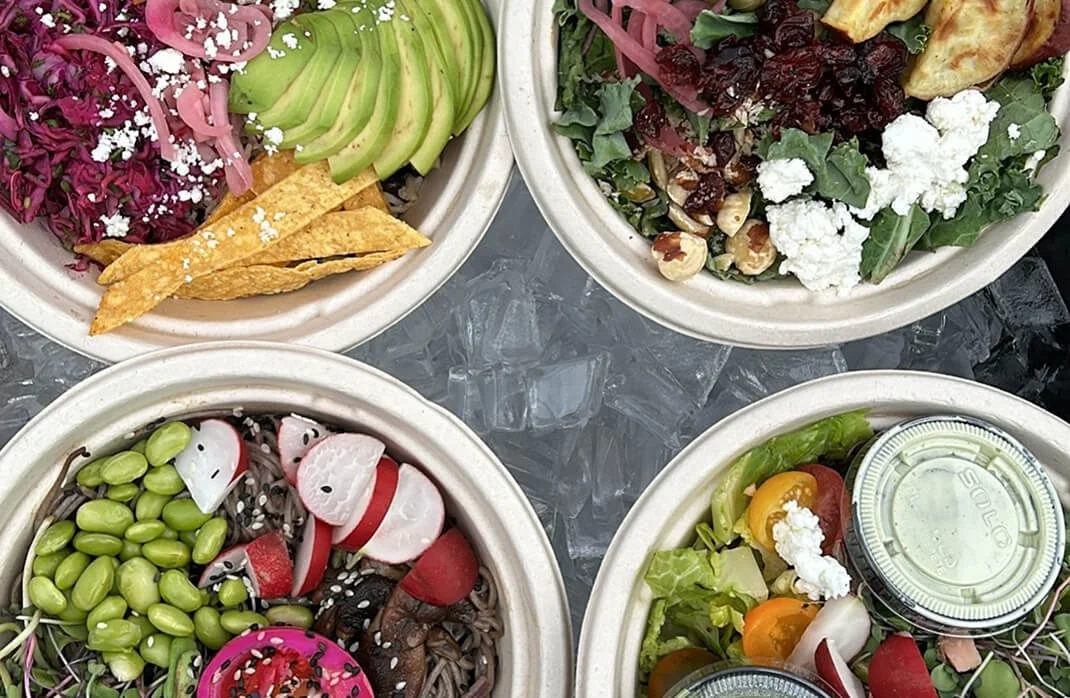 Four takeout bowls of salad with various ingredients including avocado, radishes, beans, shredded cheese, cherry tomatoes, lettuce, broccoli, and toppings like nuts, seeds, and dressings on top.