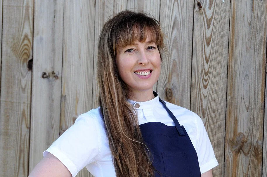 A woman with long brown hair, wearing a white shirt and navy apron, smiling in front of a wooden fence.