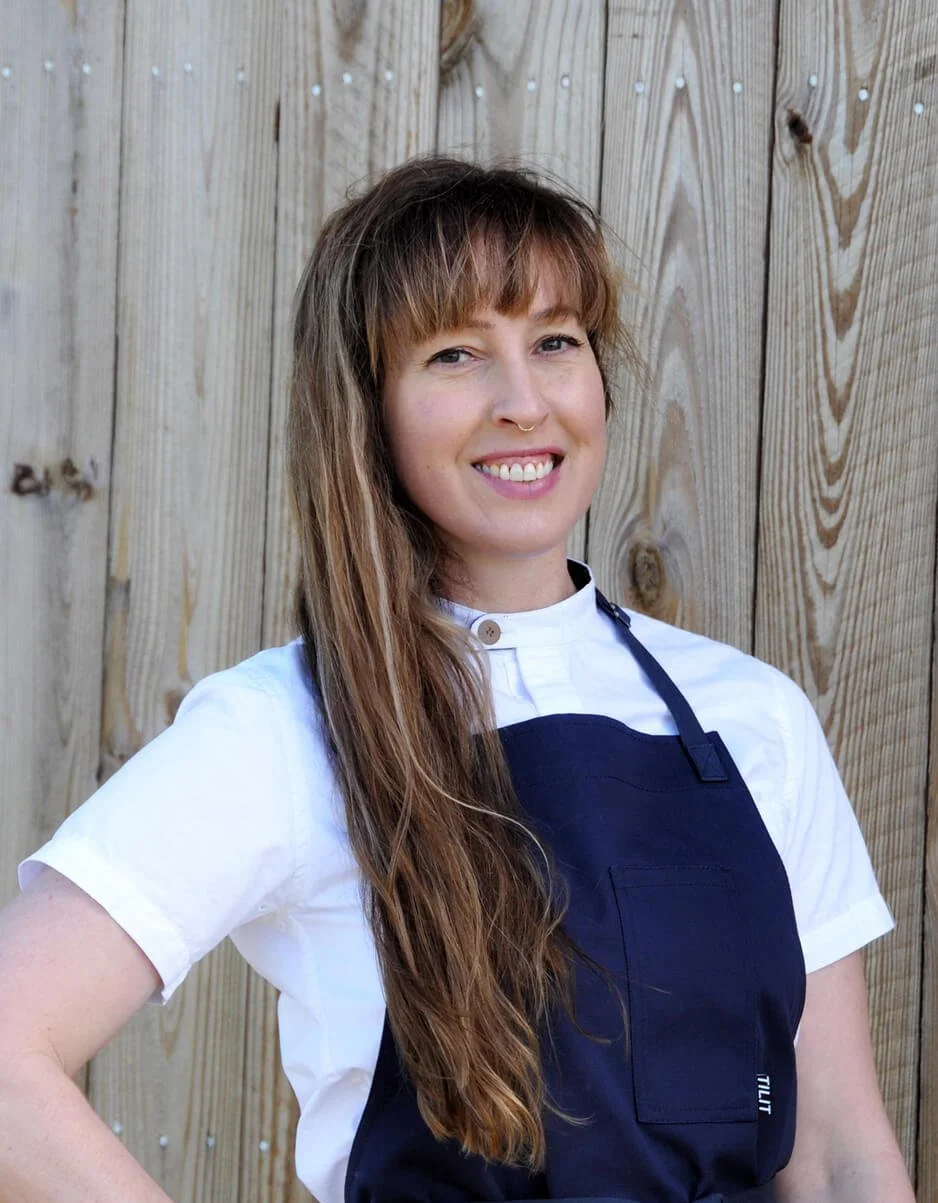 A woman with long wavy hair standing outdoors in front of a wooden fence, smiling, wearing a white shirt and a dark apron.