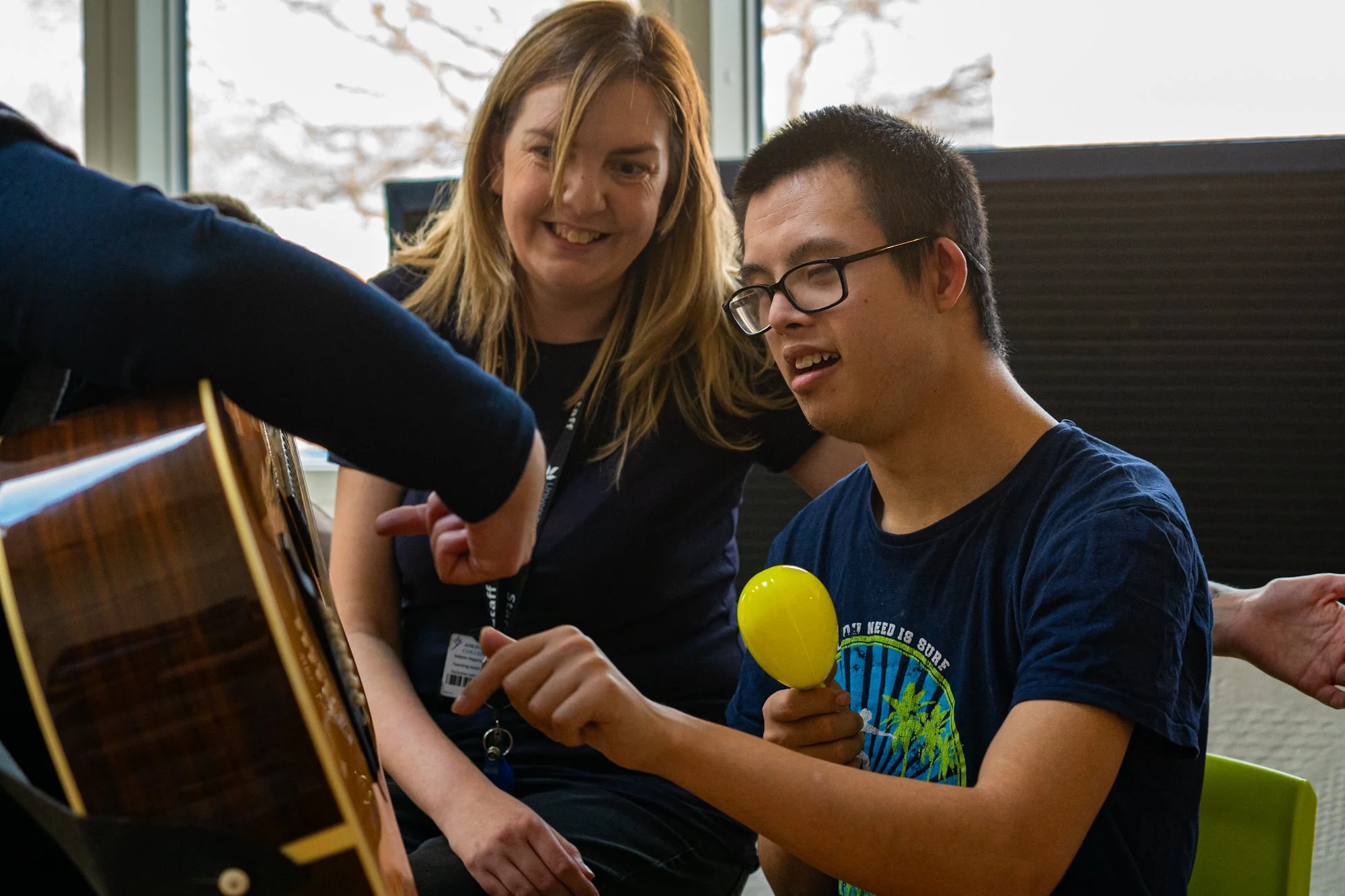 A young man wearing glasses and a blue t-shirt is sitting and playing a yellow hand drum, while a woman with blonde hair and a dark shirt looks on and smiles, inside a room with large windows showing a snowy landscape outside.