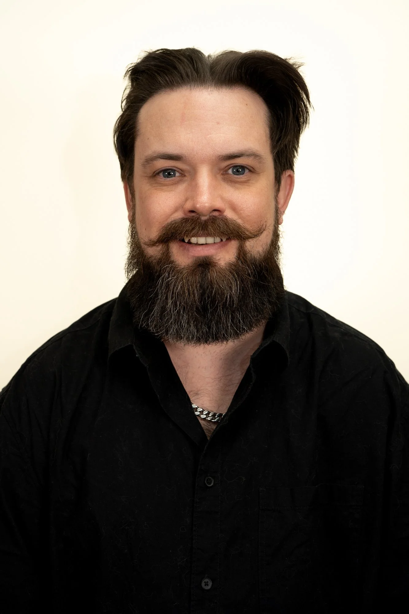 Portrait of a man with dark hair and a full beard and mustache, wearing a black shirt and a silver chain necklace, smiling against a plain white background.