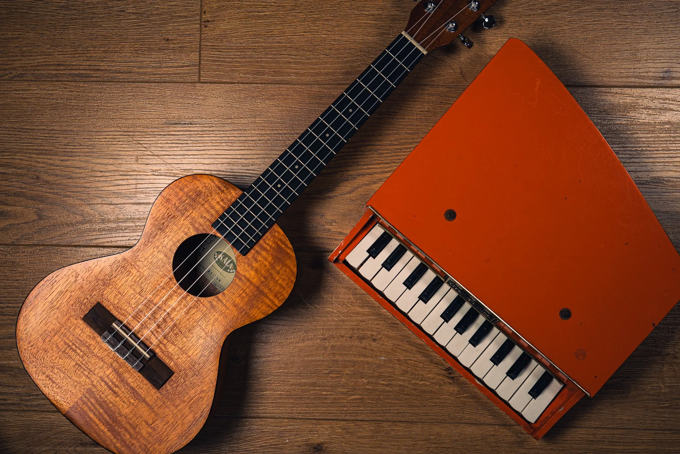 An acoustic guitar and an orange portable keyboard lying on a wooden floor.