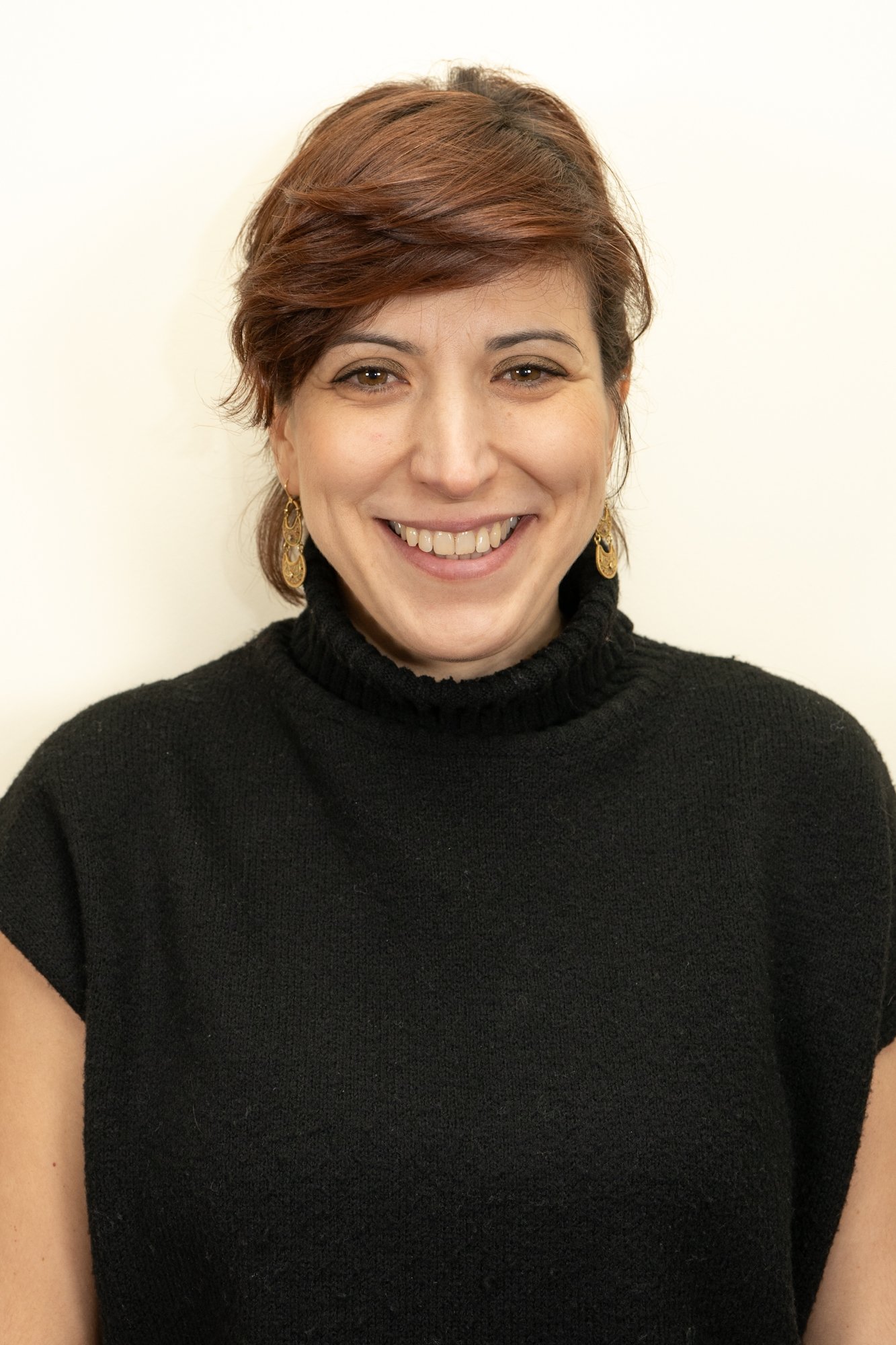 Portrait of a woman with short brown hair, wearing a black turtleneck sweater and gold earrings, smiling against a white background.