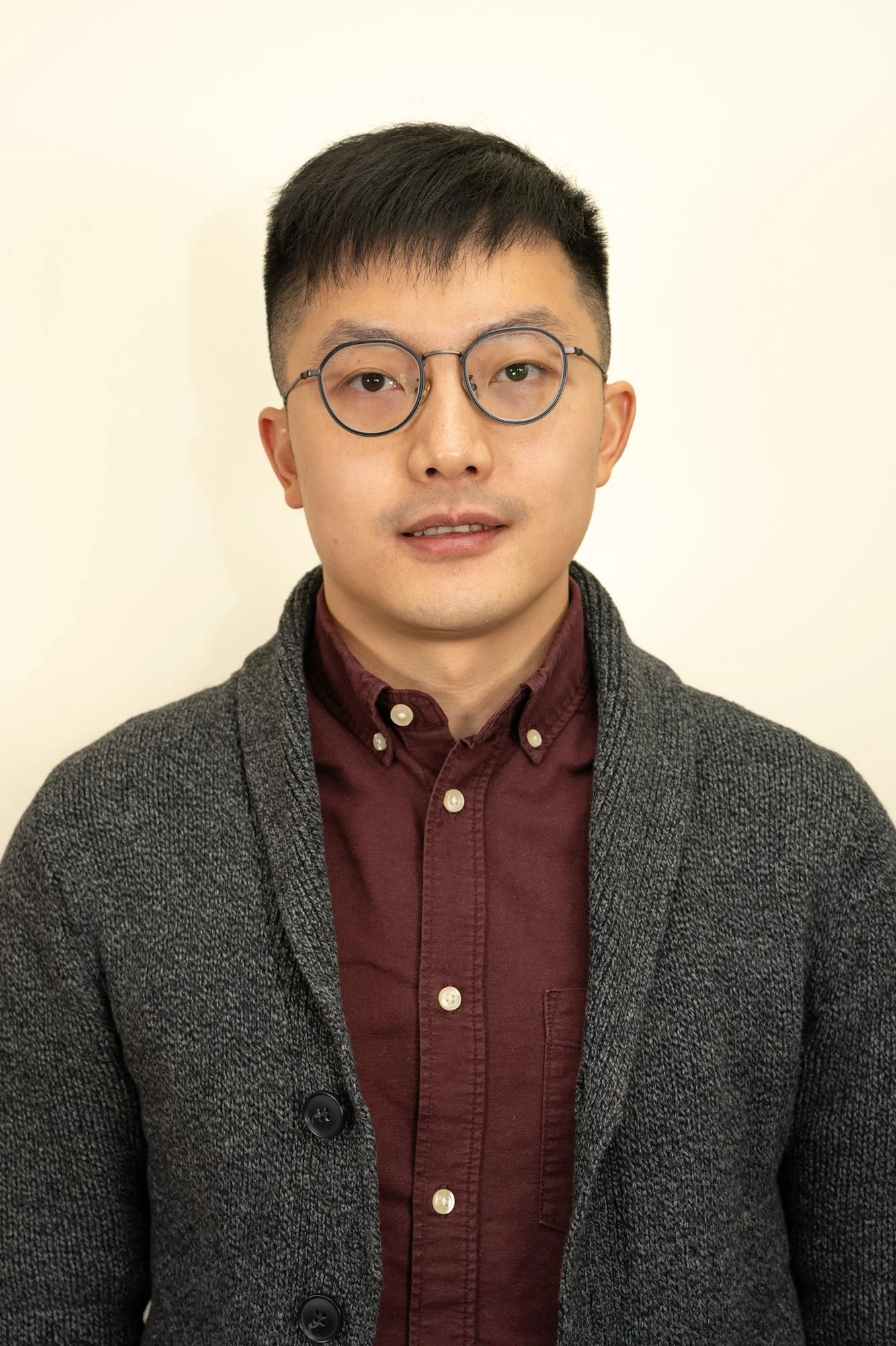 A man with black hair, glasses, wearing a dark red shirt and a gray jacket, posing against a plain off-white background.