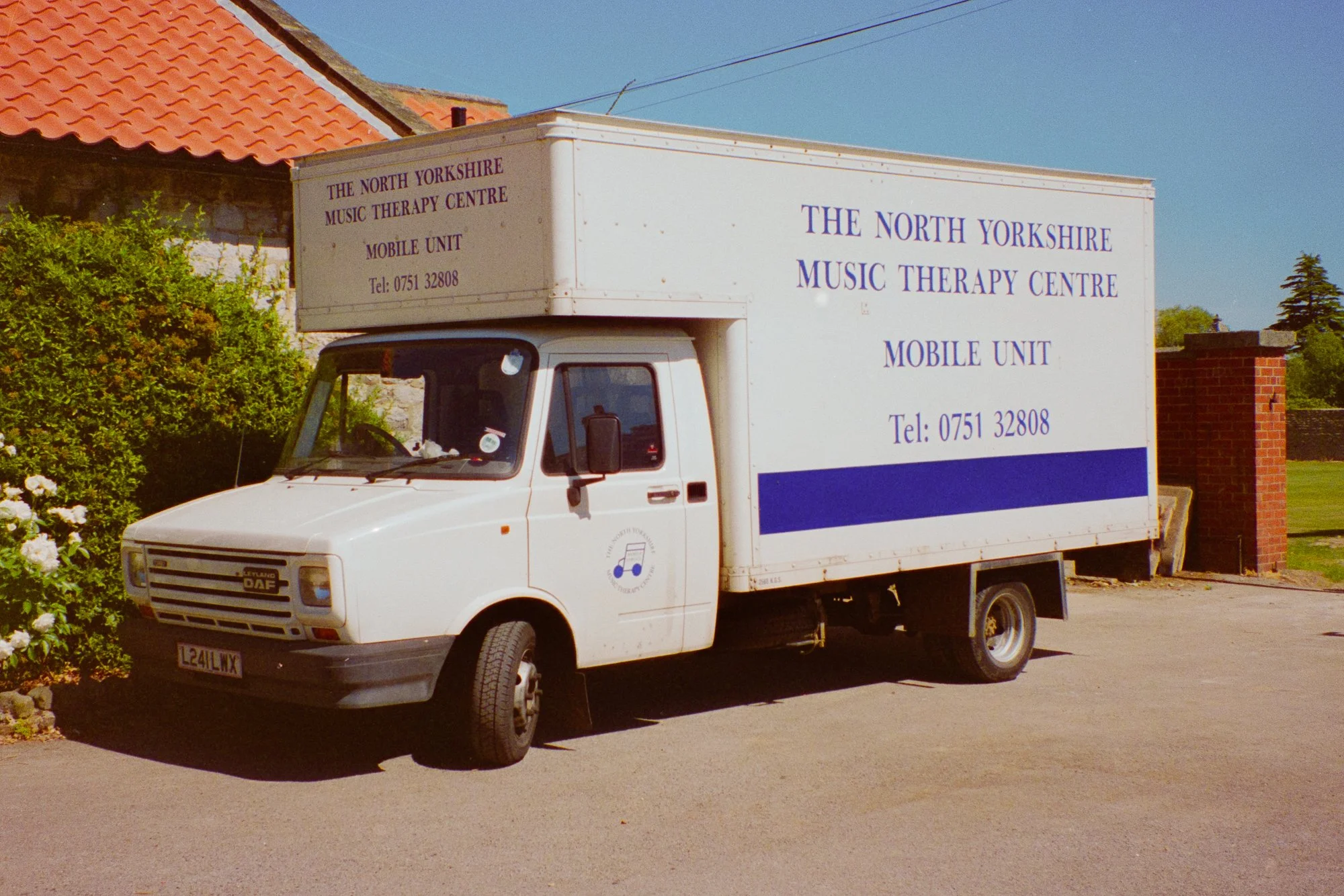 A white mobile truck with purple text on the side reading 'The North Yorkshire Music Therapy Centre' and a phone number. The truck is parked on a street next to greenery and houses with red-tiled roofs.