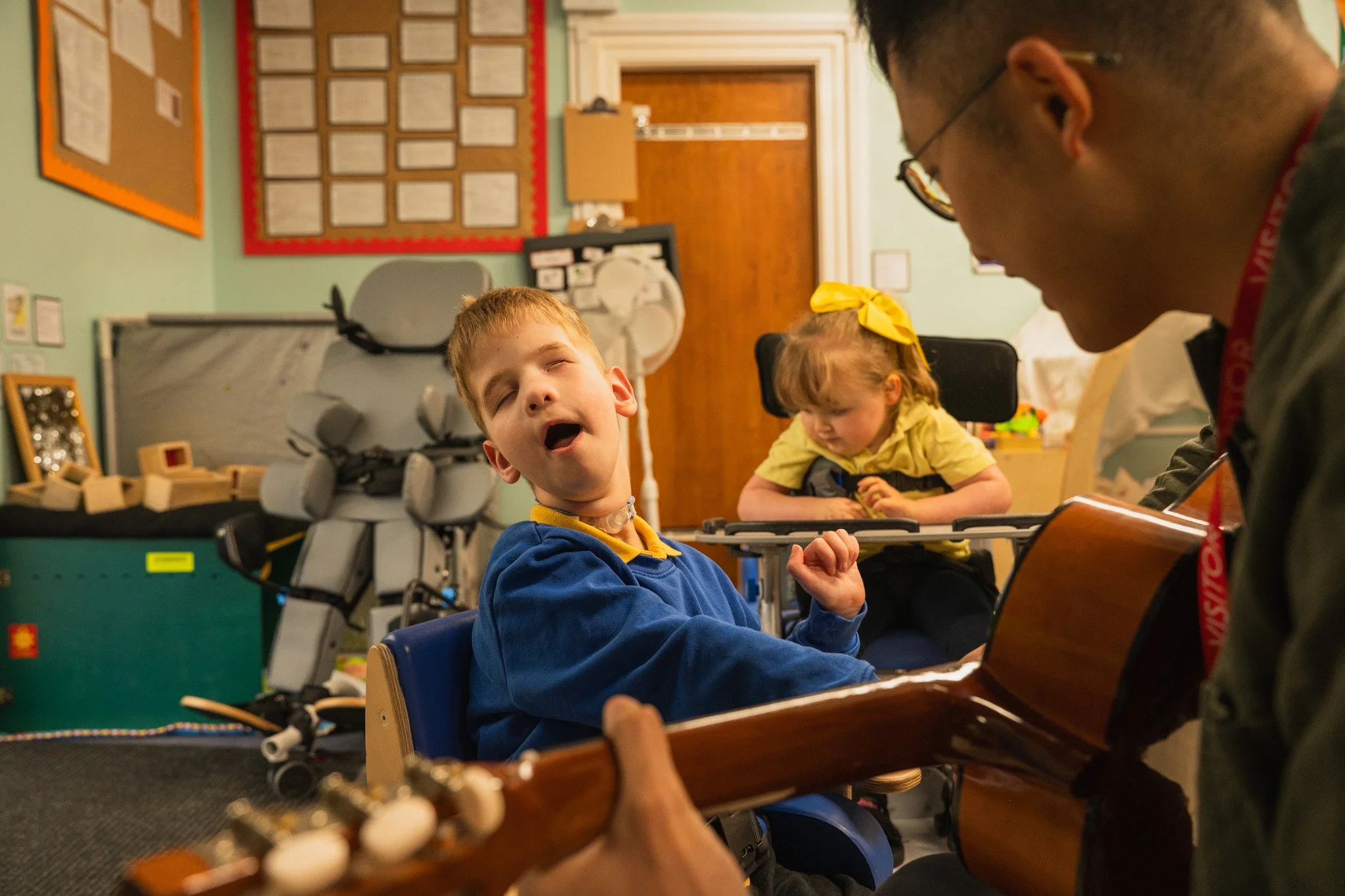 A young boy singing with his eyes closed and mouth open, sitting at a table in a classroom, while another child and a man playing guitar sit nearby. The classroom has bulletin boards, a wooden door, and various educational materials on the walls.