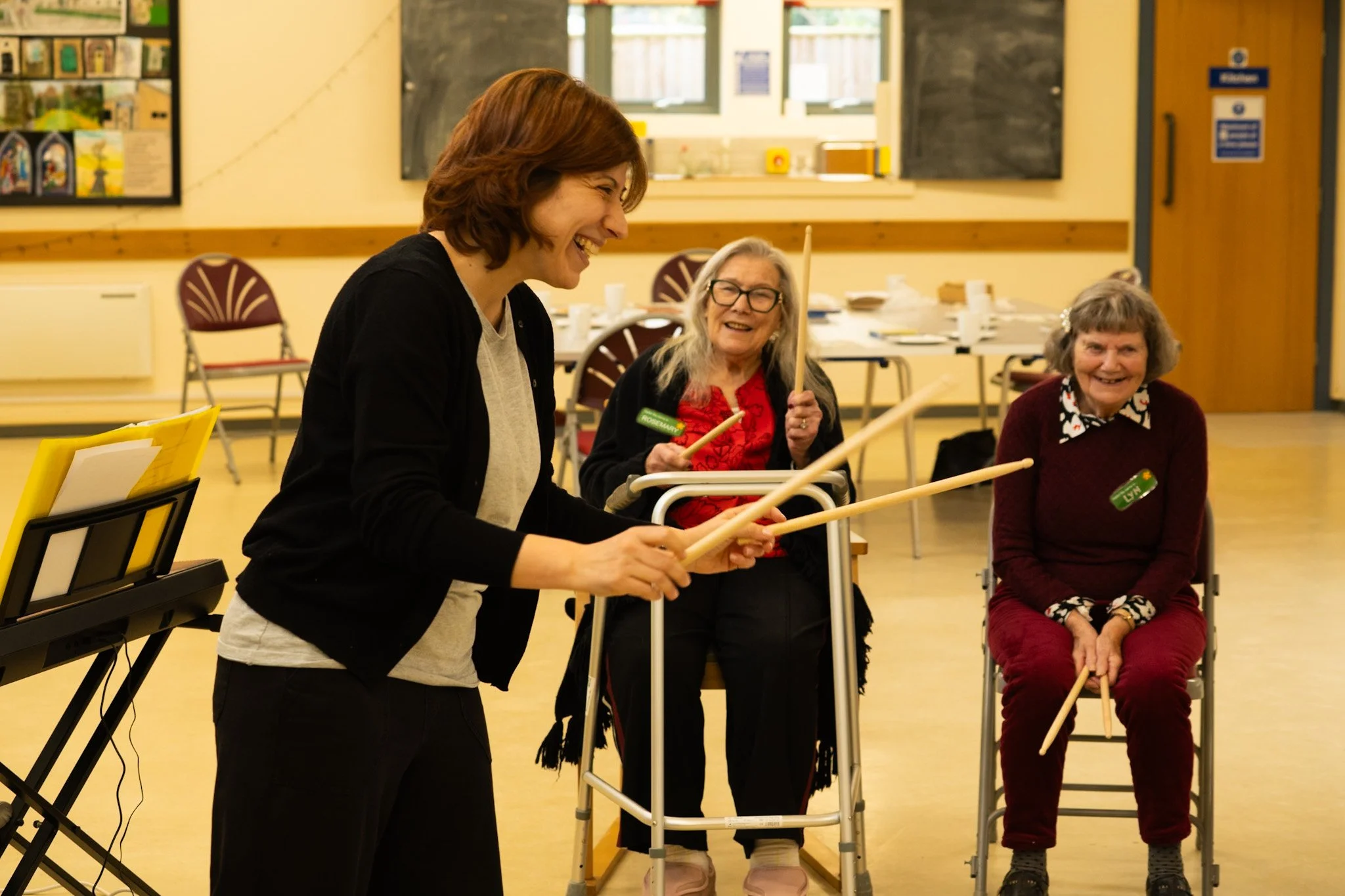 A group of elderly women seated with assistive devices, smiling and holding drumsticks, while a woman with red hair standing and playing a keyboard.