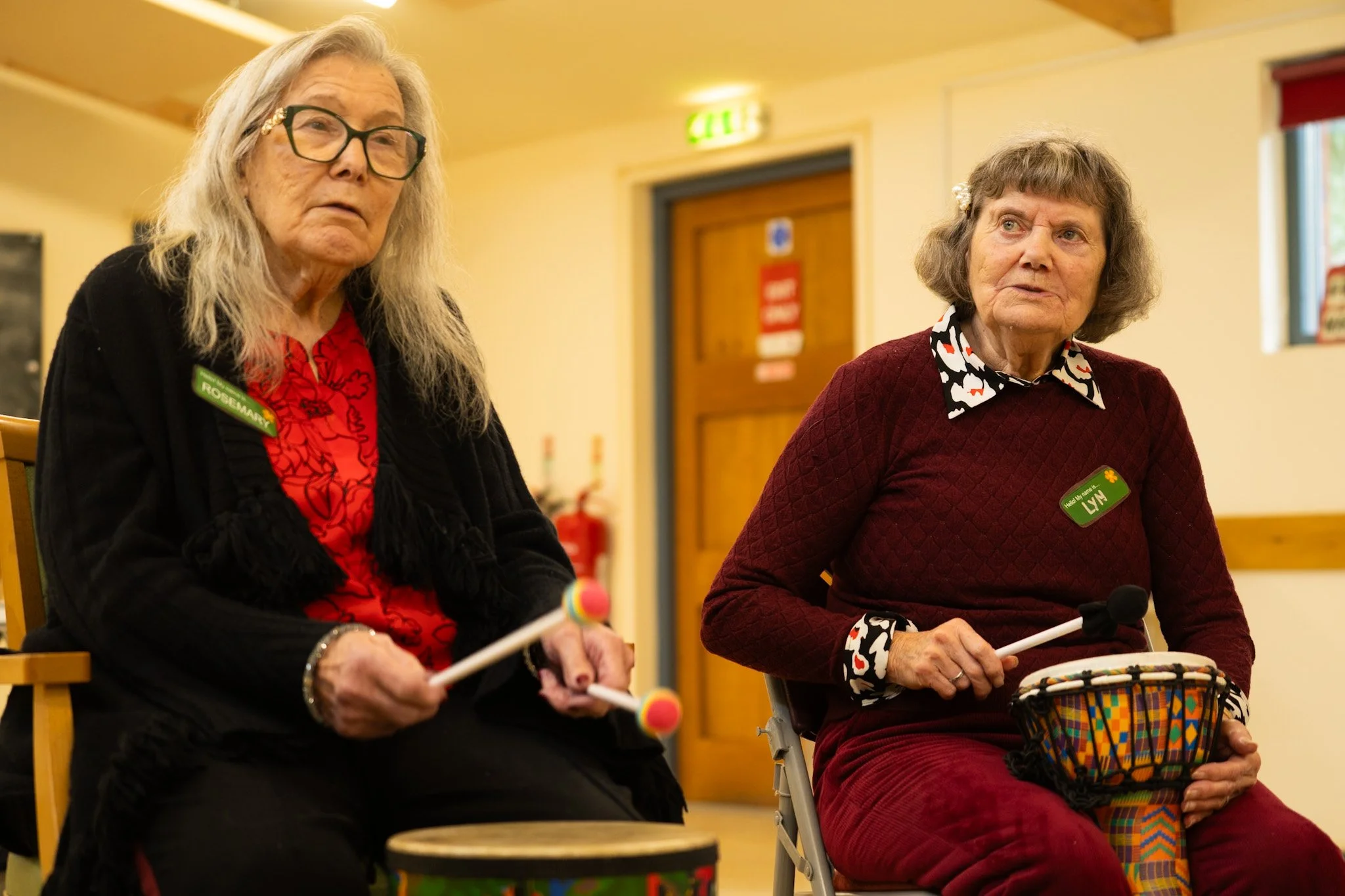 Two elderly women sitting indoors, playing musical instruments, with a yellow wall, a wooden door, and a window in the background.