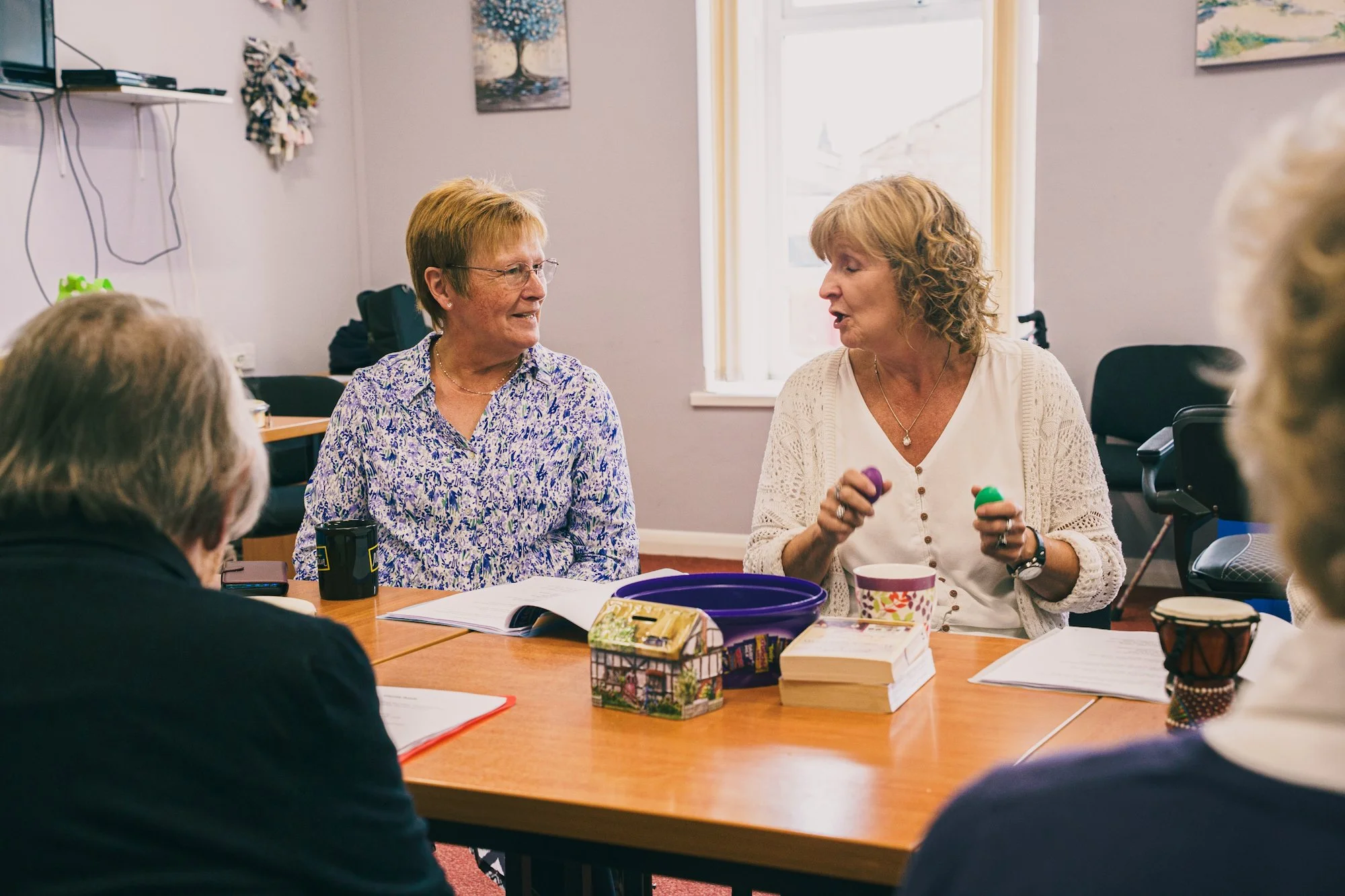 A group of older adults sitting around a table in a well-lit room, engaged in conversation. Two women are prominent in the center, one with short hair and glasses wearing a floral shirt, and another with curly hair in a white top holding colorful objects.