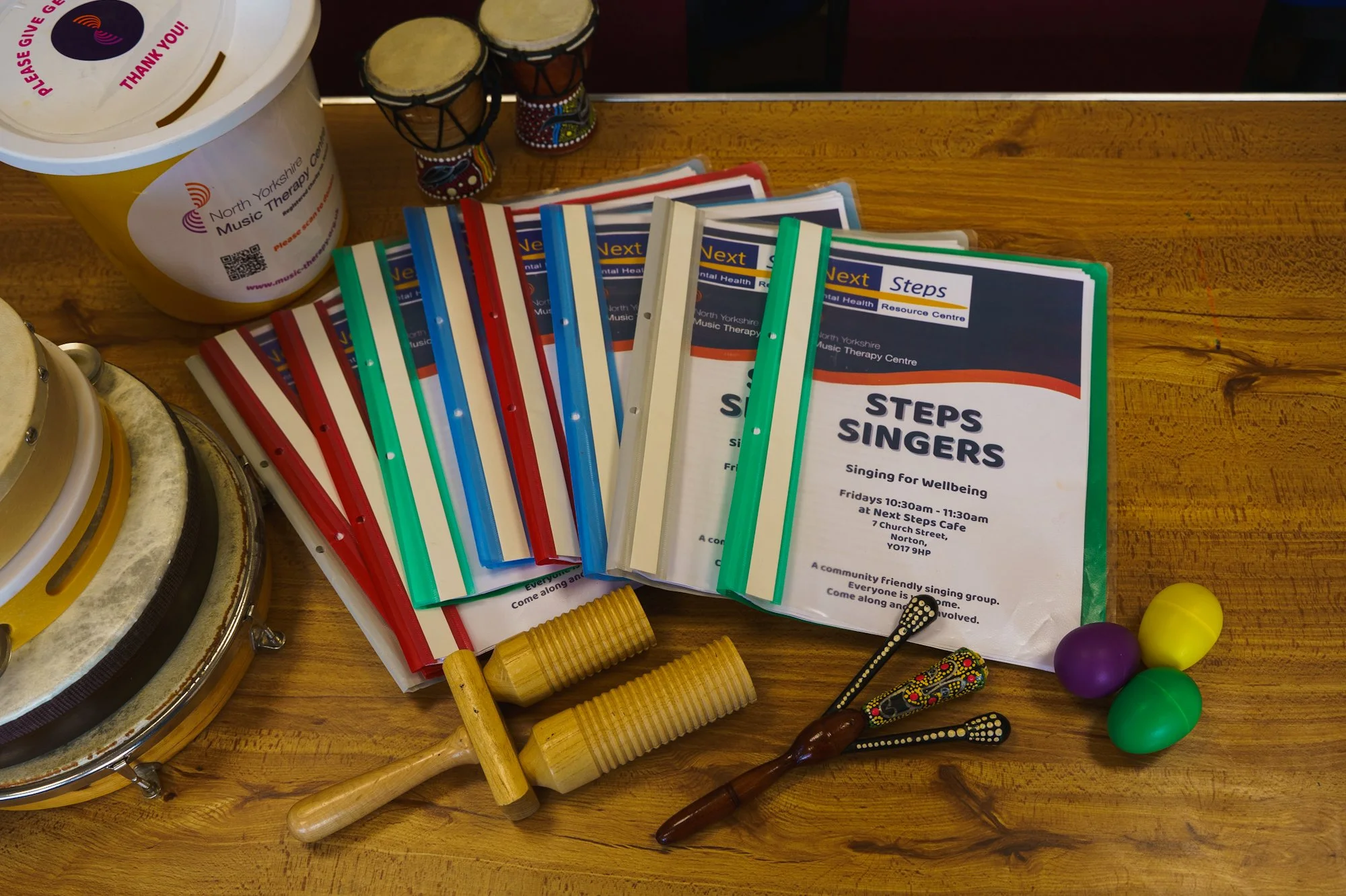 A wooden table with various objects, including colorful printed booklets titled 'Next Steps SINGERS', two small decorative drums, a yellow container labeled 'North Yorkshire Music Therapy Centre', and some percussion instruments like tambourines, maracas, and a wooden frog-shaped shaker.
