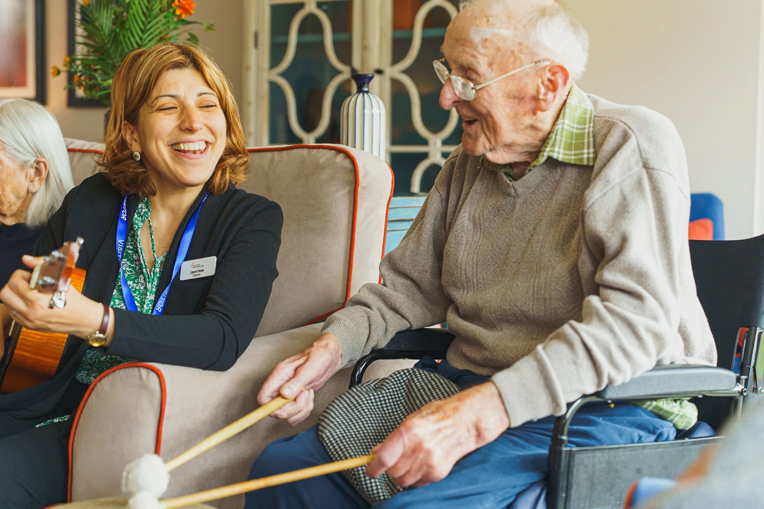 A young woman with red hair and a name tag playing guitar and smiling at an elderly man in a wheelchair who is holding her hand and smiling back, with another elderly person partially visible on the left side of the image.