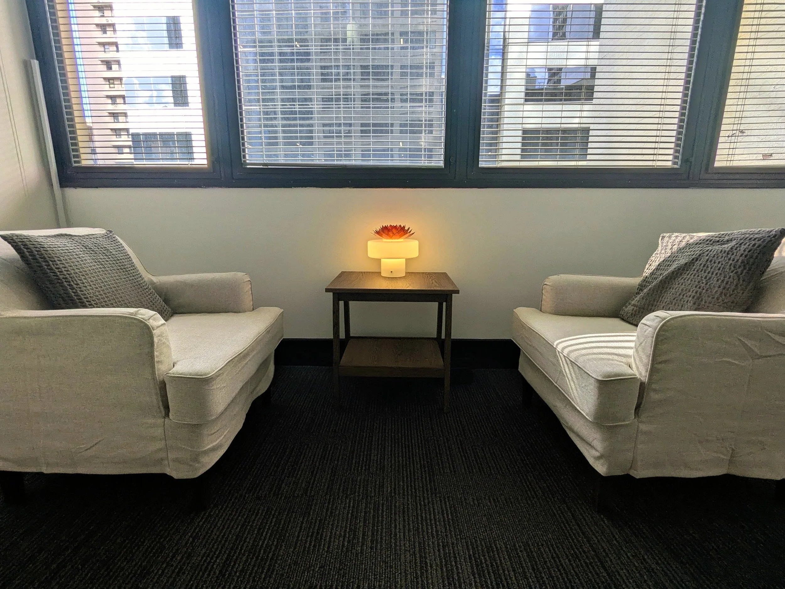 A counselling room with two comfortable armchairs facing each other, lamp with a warm glow on a wooden coffee table in the center, with a window, promoting a relaxing, safe and comforting place to share your stories