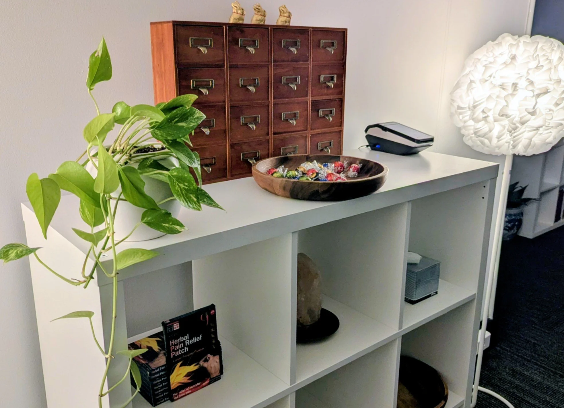 A white shelf with a potted green plant, a wooden catalog box symbolising eastern medicine herbal drawers, a large white paper lantern lamp - demonstrating balanced calm, practicality and vitality
