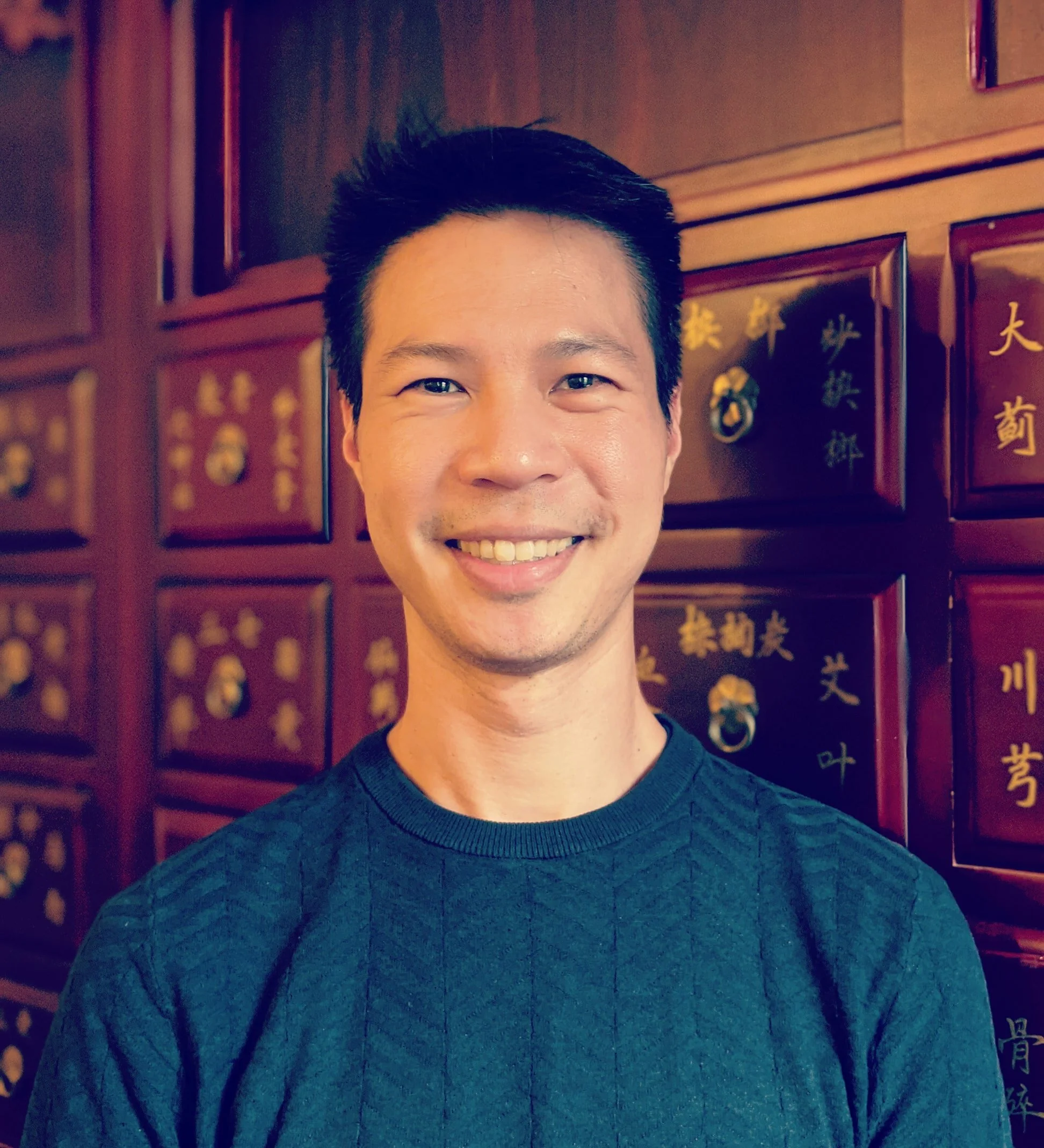 portrait photo of Francis, a cis-man of South East Asian background, who is wearing a dark coloured shirt, has short, dark hair and has a welcoming smile on his face