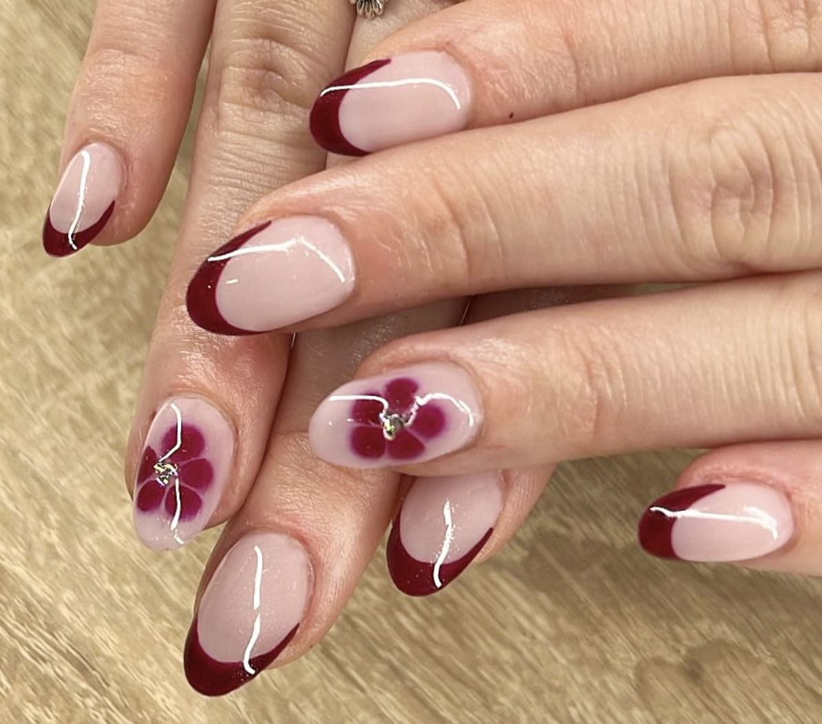 On voit des ongles manucurés avec un design en French manicure, en couleur bordeaux sur le bord des ongles, et avec un motif en forme de fleur rouge sur certains ongles.