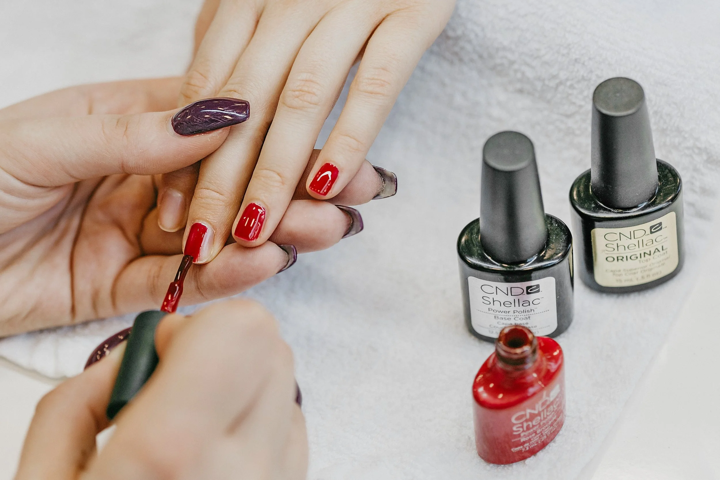 Une personne appliquant du vernis à ongles rouge sur ses ongles, avec plusieurs vernis à ongles en bouteille sur une surface. La personne a déjà un vernis à ongles violet. Des bouteilles de vernis à ongles de la marque CND Shellac sont visibles.