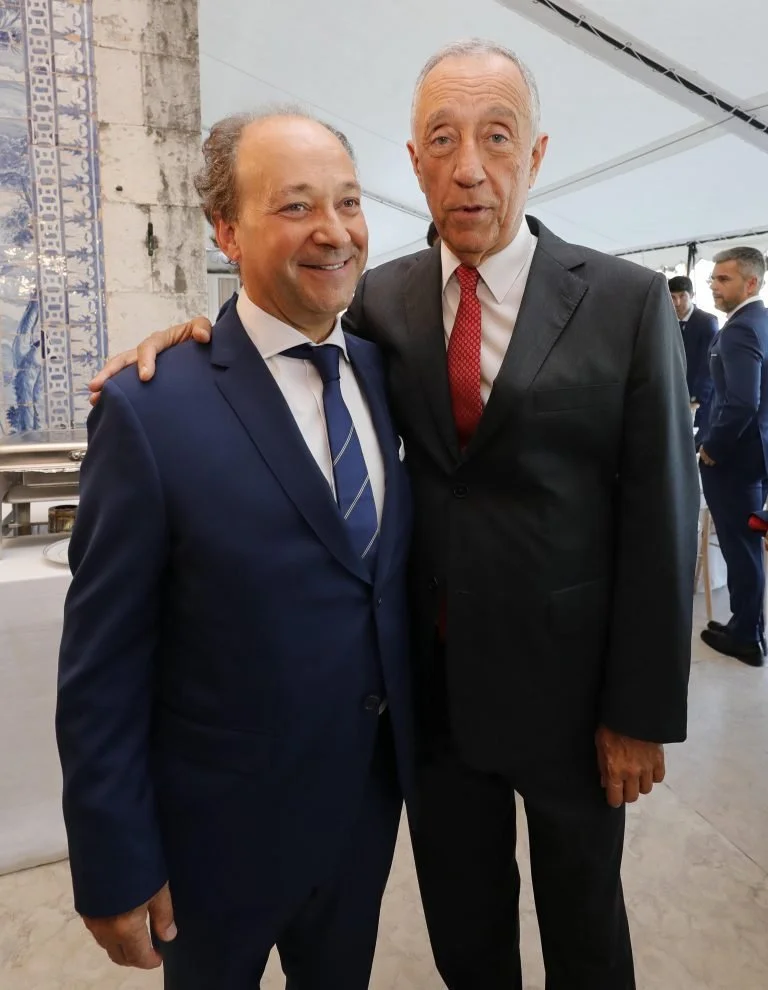 Dois homens vestidos com terno, sorrindo e posando lado a lado em um evento formal, um deles com braço no ombro do outro.