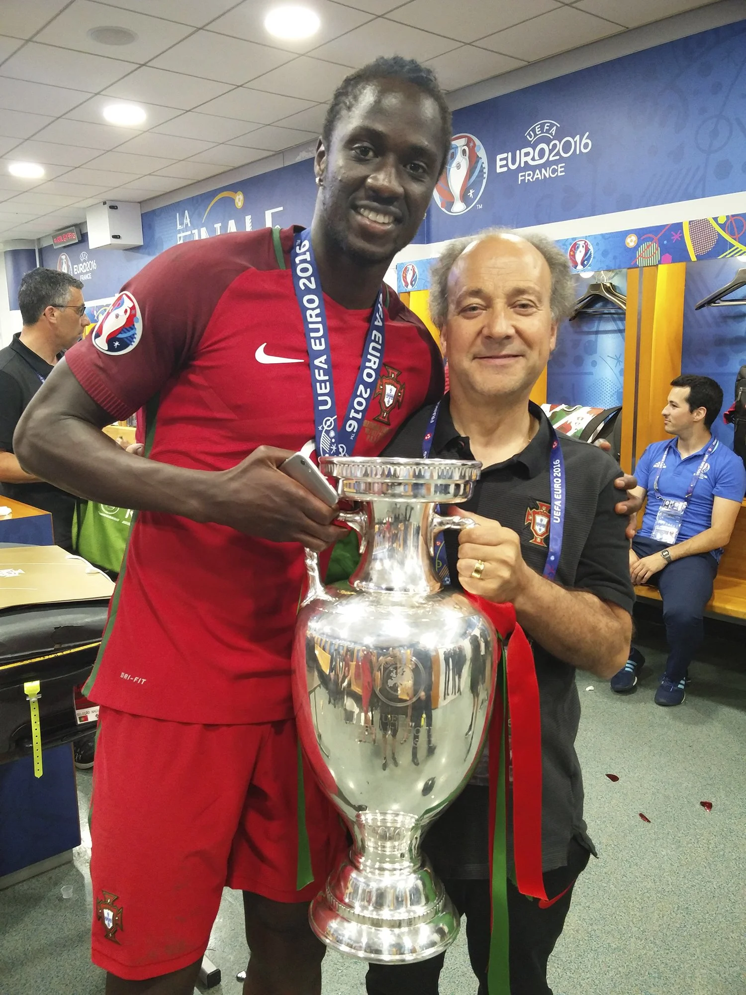 Dois homens comemorando com uma taça de campeonato, um dos homens usa uniforme de futebol da seleção de Portugal e medalha de UEFA Euro 2016, o outro homem segura a taça e aponta para ela.