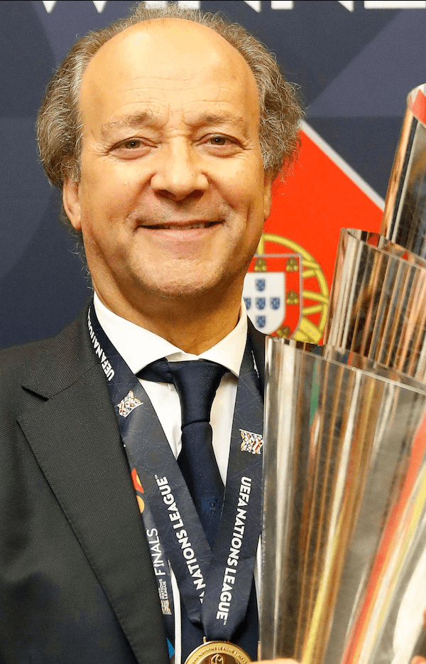 Homem de terno sorrindo, usando uma medalha do UEFA Champions League e segurando uma taça de troféu, com fundo mostrando o escudo de Portugal.