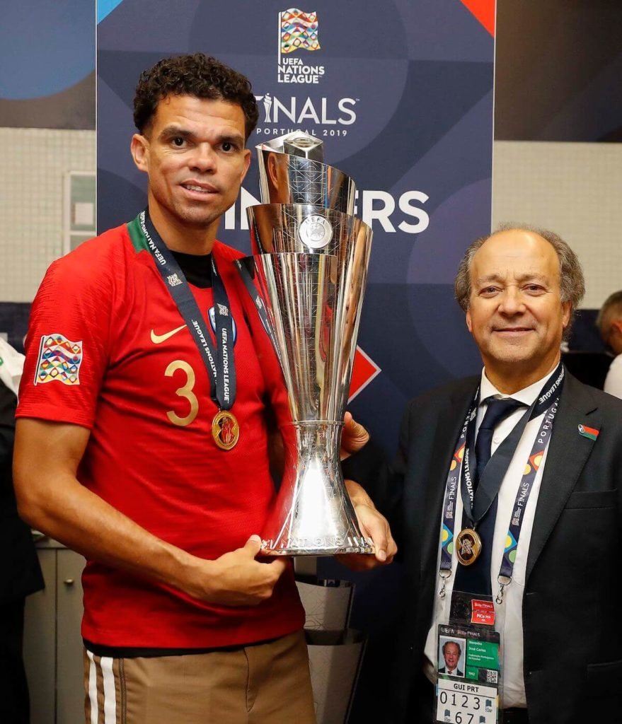 Dois homens celebram com um troféu da Liga das Nações da UEFA em um evento, um usando uma camisa vermelha com o número 3 e uma medalha, o outro vestindo terno e crachá de credencial, ambos sorrindo ao lado do troféu.