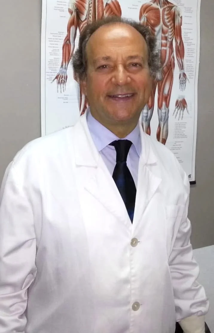 Homem sorridente usando jaleco branco e gravata em consultório com poster anatômico das musculaturas do corpo humano ao fundo.