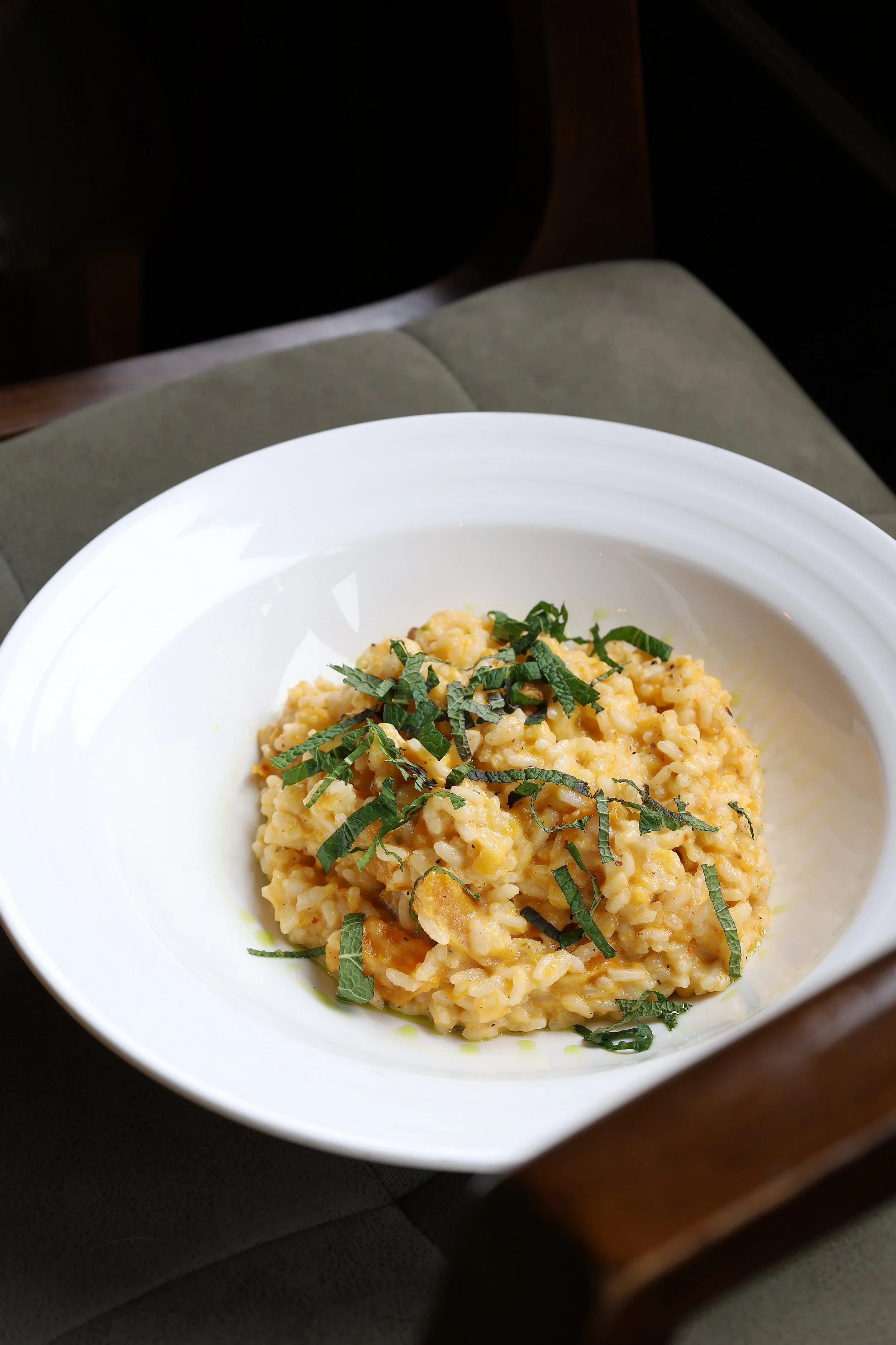 Close-up of a bowl of creamy risotto garnished with chopped herbs, sitting on a dark surface with a bed and furniture in the background.