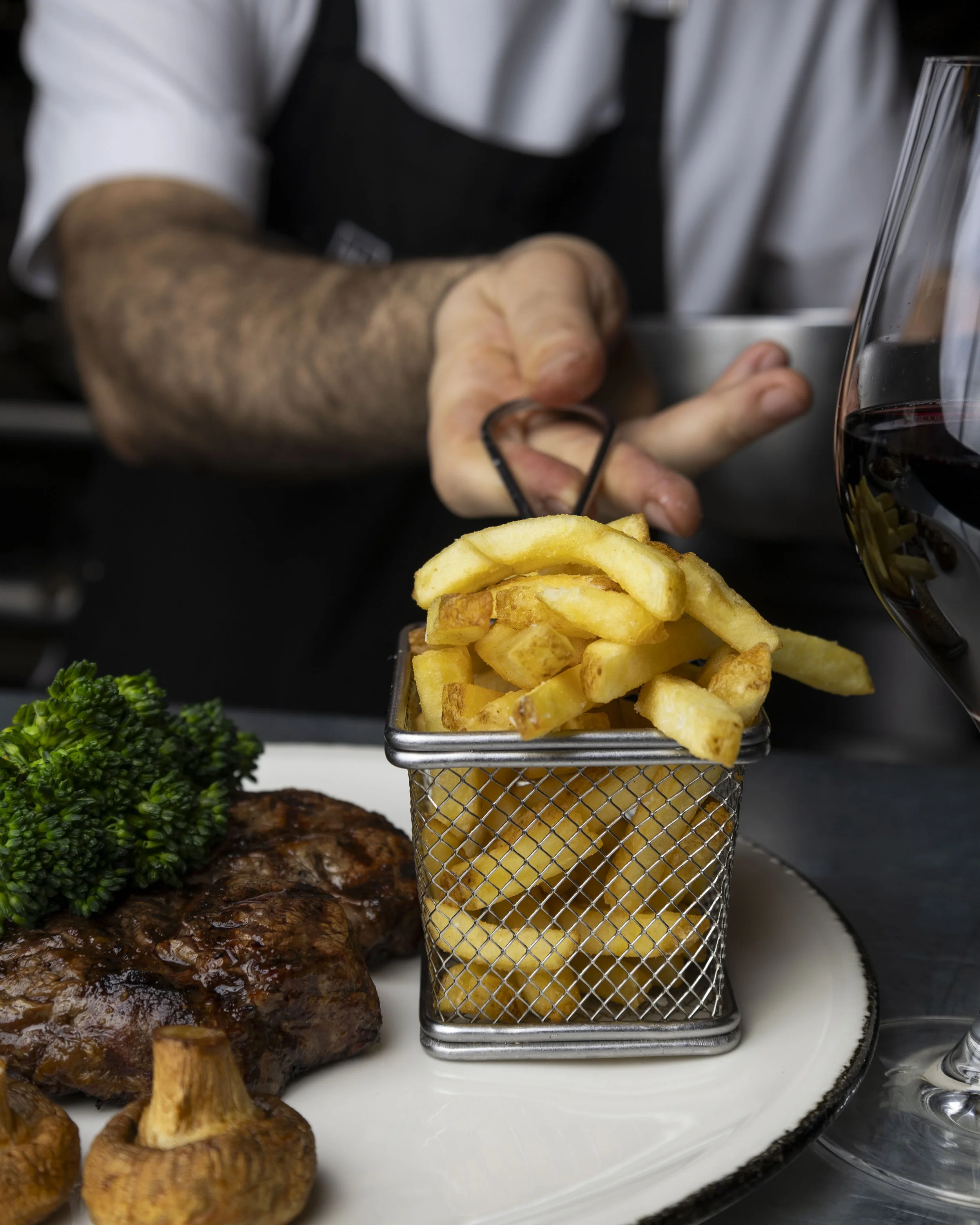 Close-up of a restaurant meal with French fries in a small metal basket, grilled chicken with sauce, sautéed mushrooms, broccoli, and a glass of red wine on a white plate
