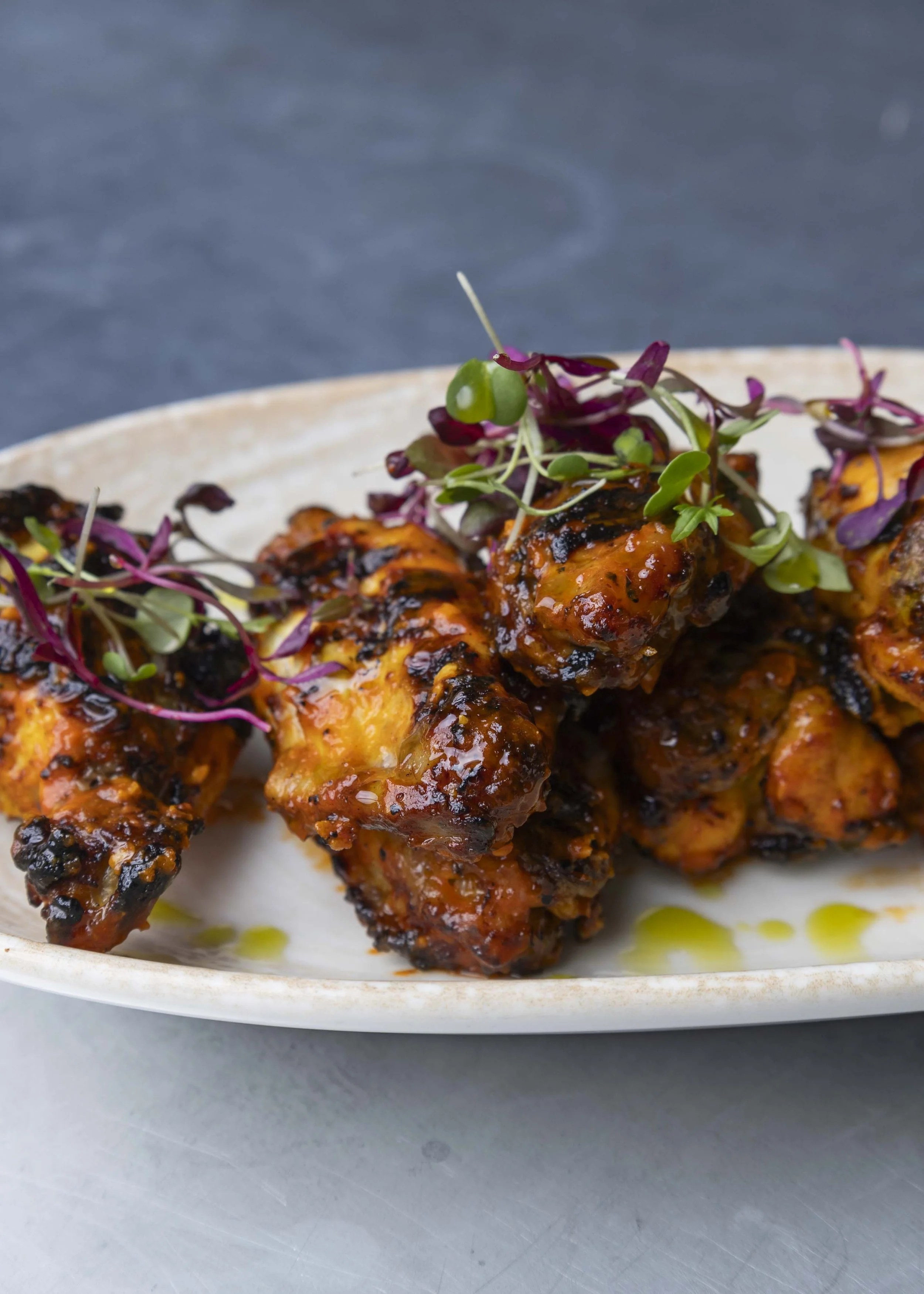 Close-up of grilled chicken wings coated in a spicy sauce, garnished with microgreens, served in a white ceramic dish on a gray surface.