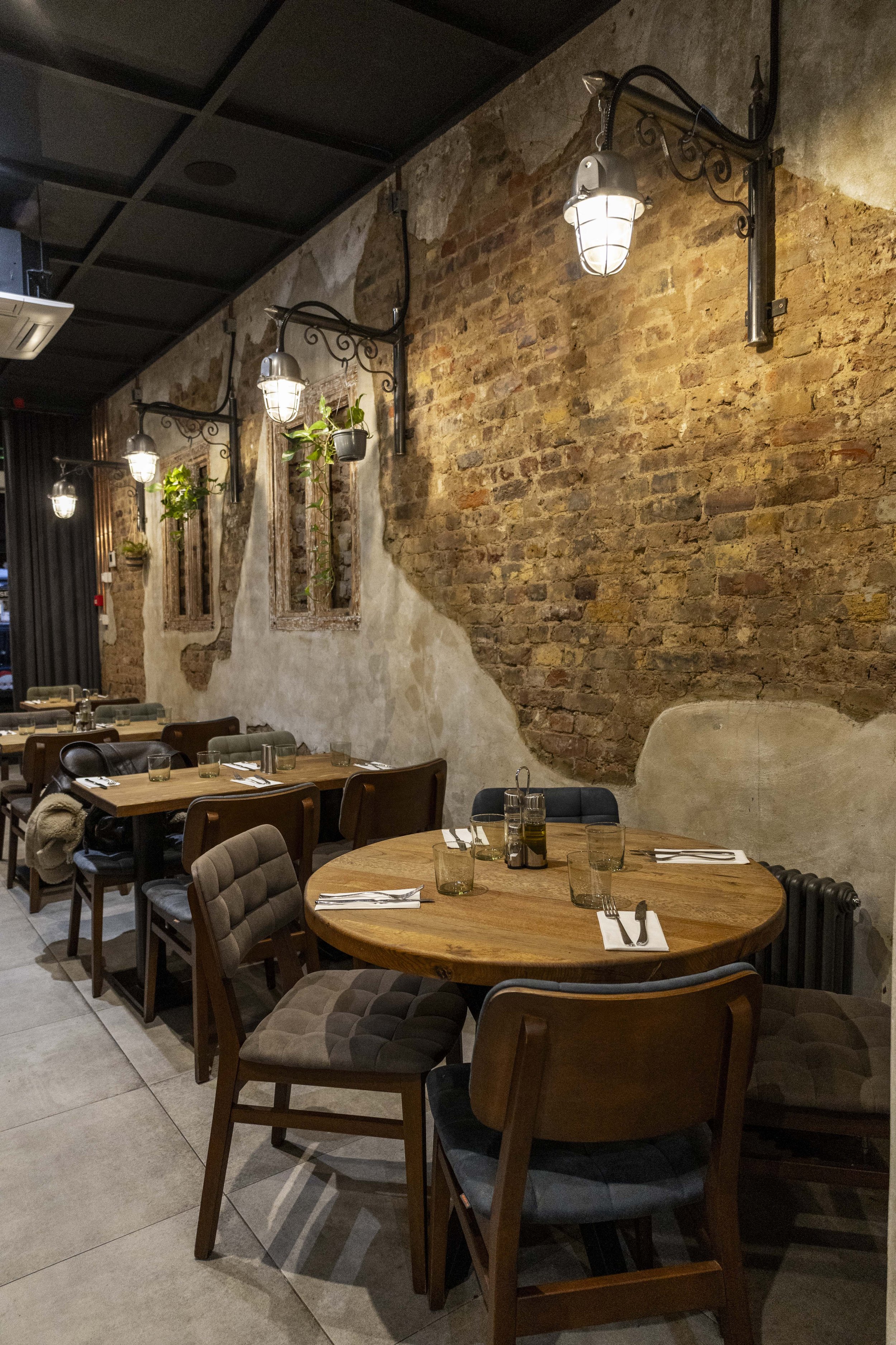 Interior of a restaurant with wooden tables and assorted chairs, exposed brick and plaster walls, vintage wall-mounted lamps, and hanging potted plants.