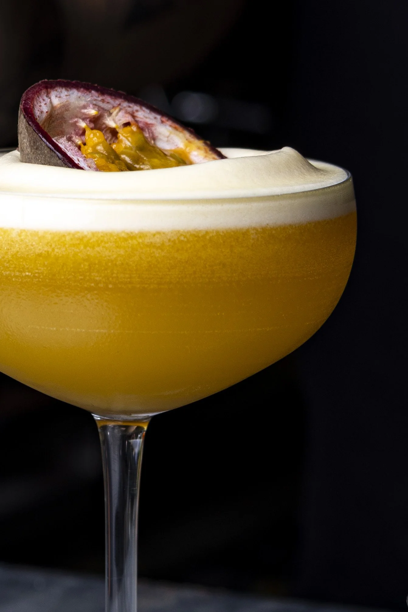 A cocktail glass filled with a yellow beverage, topped with whipped cream and garnished with a halved passion fruit.