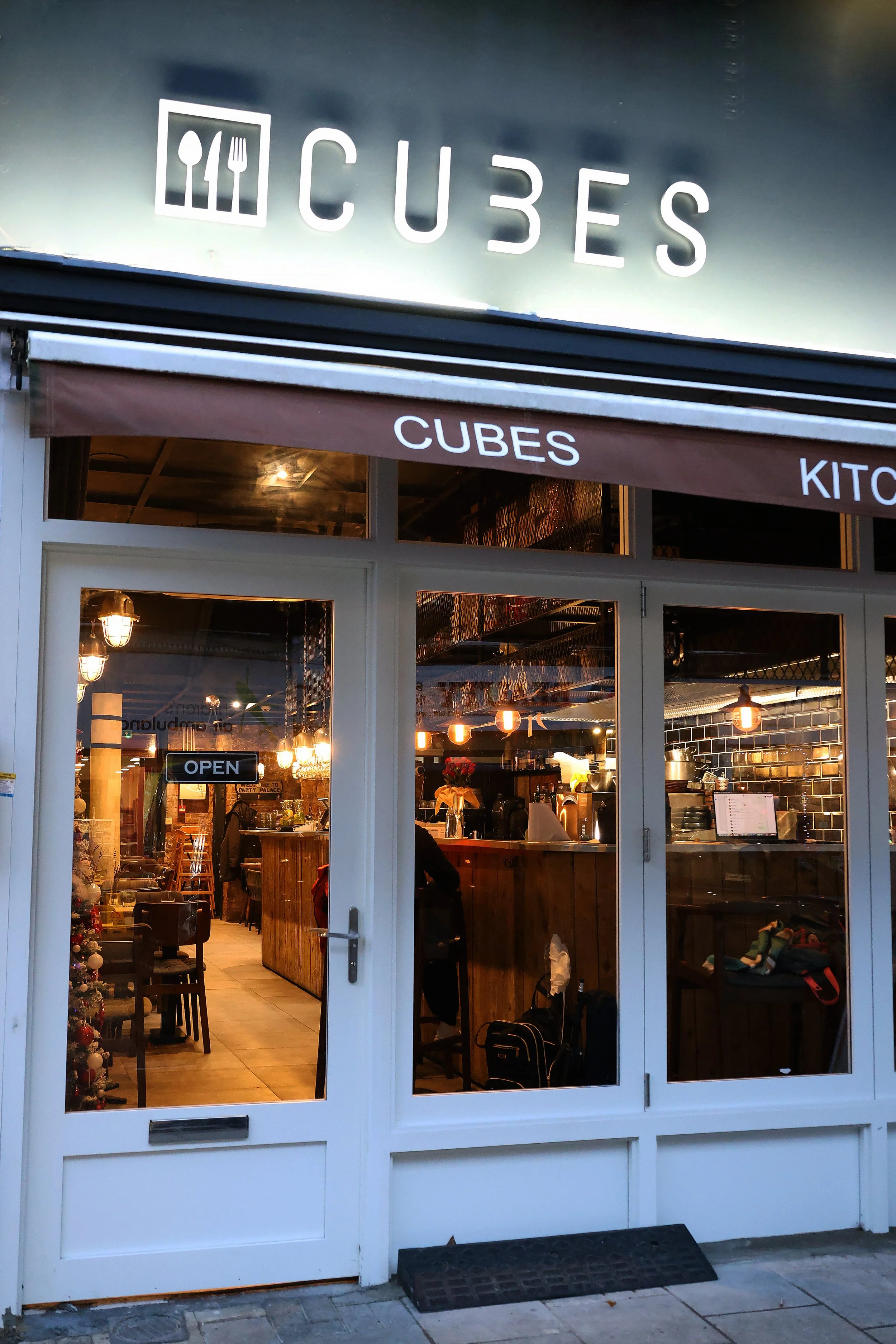 A cafe or restaurant from the outside with large glass windows and a door. The shop has a sign on top that reads 'CUBES' with an icon of a spoon, fork, and knife in a square. Inside, various lighting fixtures and furniture are visible, along with som