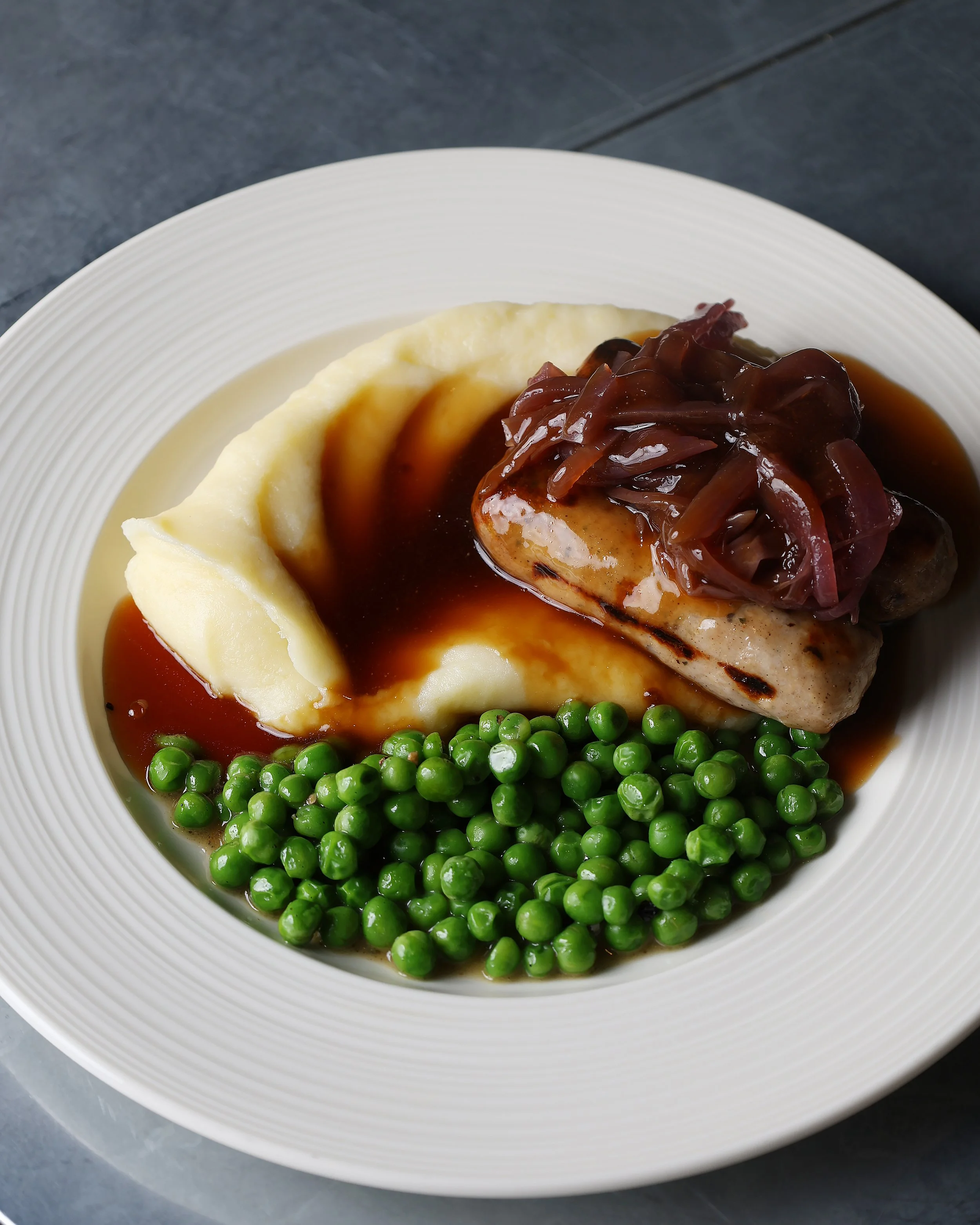 A white plate with mashed potatoes topped with brown gravy, green peas, and a portion of meatloaf topped with caramelized onions.