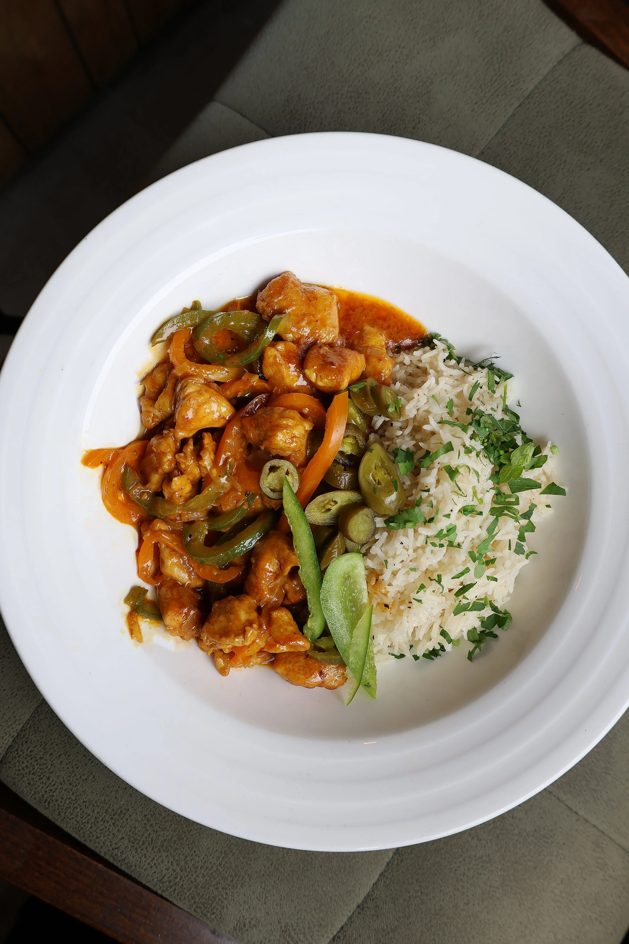 A white plate of Asian food with rice garnished with chopped green herbs, stir-fried vegetables, and pieces of chicken in orange sauce, placed on a dark wooden surface with a gray cloth underneath.