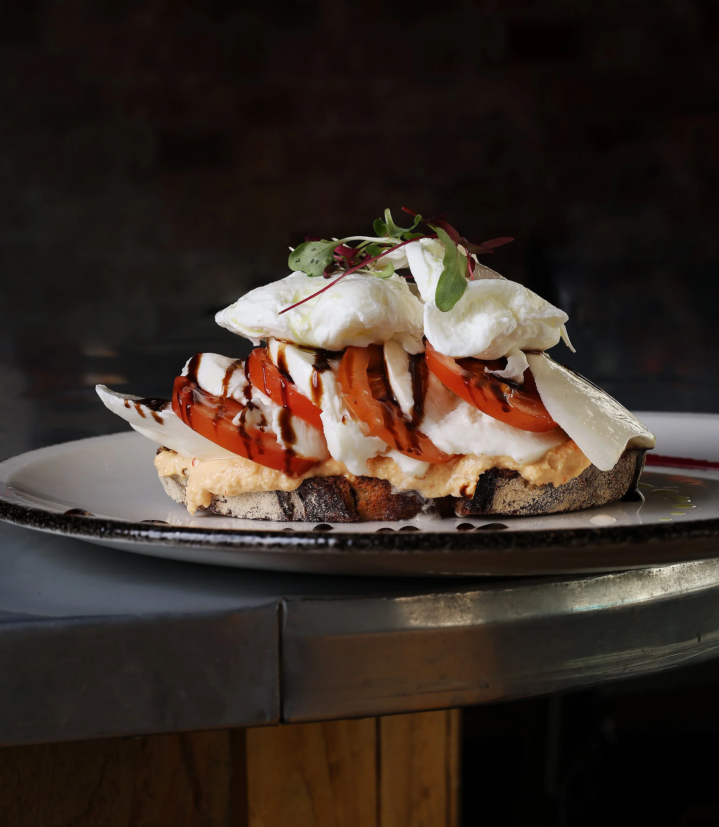 Close-up of a dessert with whipped cream, sliced strawberries, chocolate syrup, and microgreens on a black plate.