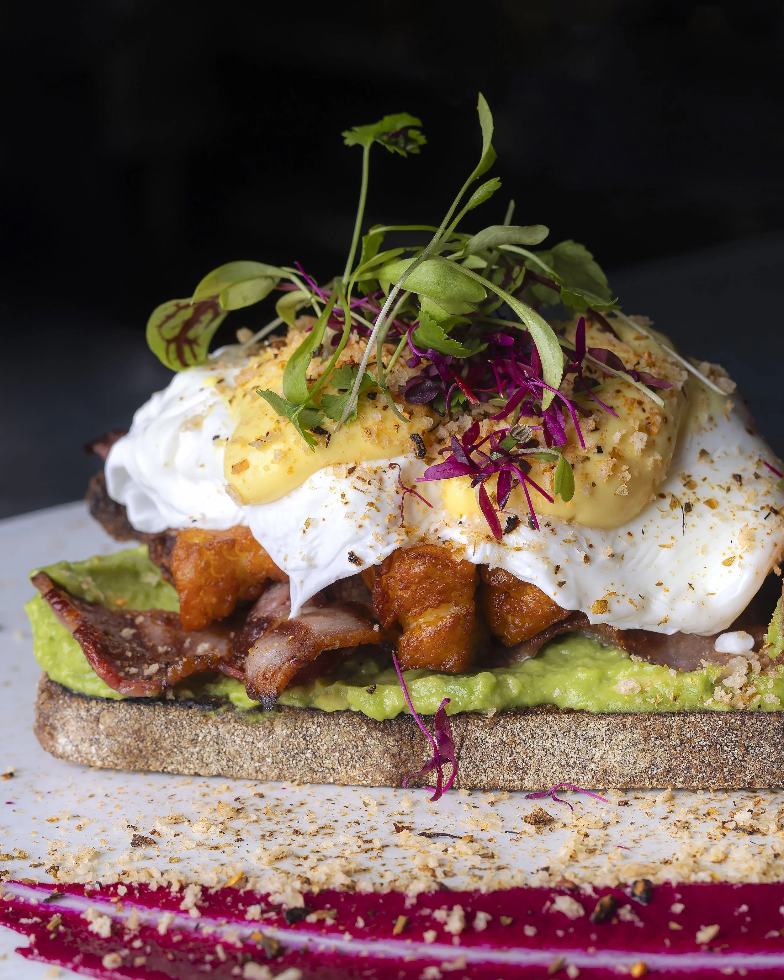 Close-up of a gourmet sandwich with layers of lettuce, crispy bacon, poached egg, microgreens, and a drizzle of sauce on a whole grain bun, with crumbs and tiny microgreens scattered around.