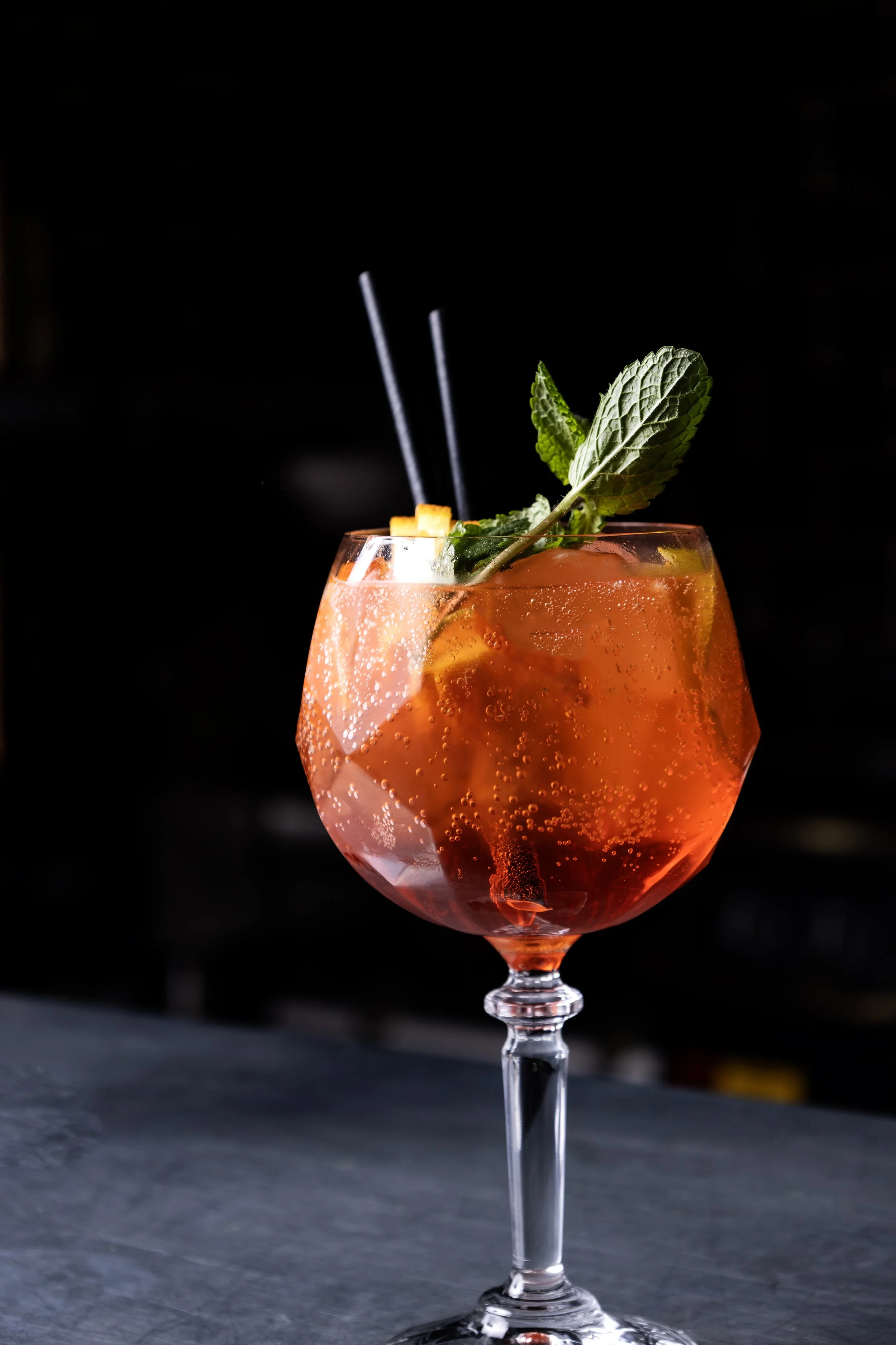 A cocktail in an elegant glass garnished with fresh mint leaves and an orange twist, with black straws, against a dark background.