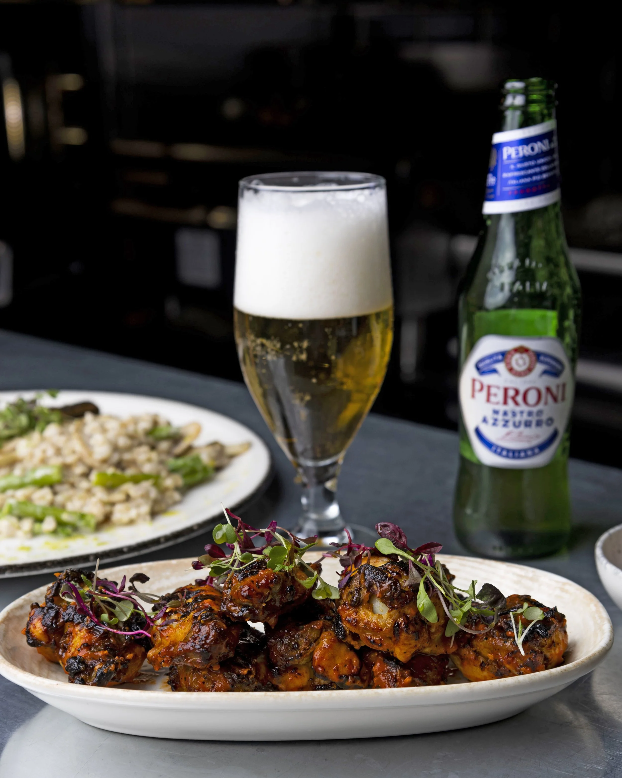 A white plate with grilled chicken pieces topped with microgreens, served on a gray surface. In the background, there is a glass of beer with frothy foam and a bottle of Peroni beer. Also, there is a partially visible dish with rice and vegetables.