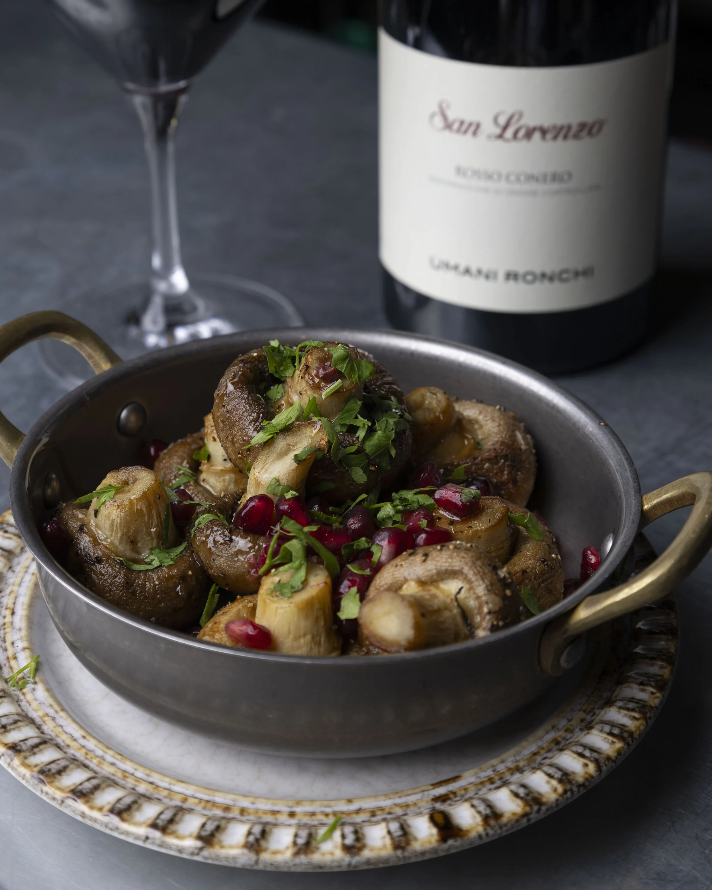 Close-up of a metal skillet containing cooked mushrooms, garnished with chopped parsley and pomegranate seeds, set on a decorative ceramic plate with a gold handle. In the background, a bottle of San Lorenzo wine and a partially visible wine glass ar