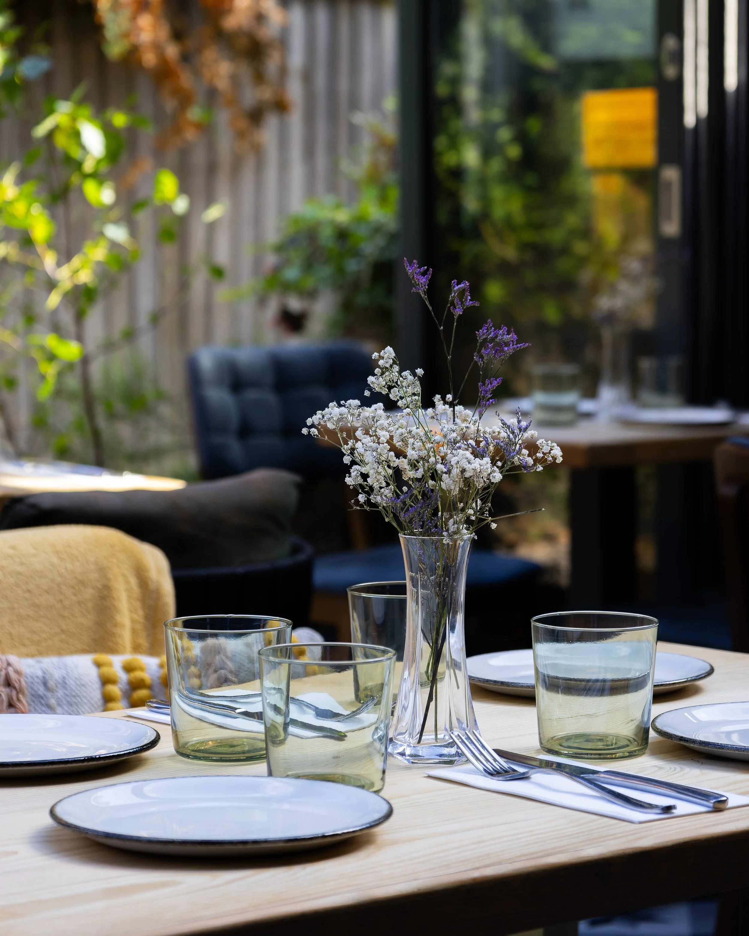A table set for a meal with white plates, clear glasses of water, and silver utensils, topped with a bouquet of white and purple flowers in a tall glass vase. The background shows a cozy outdoor space with a wooden fence, greenery, and a black chair.