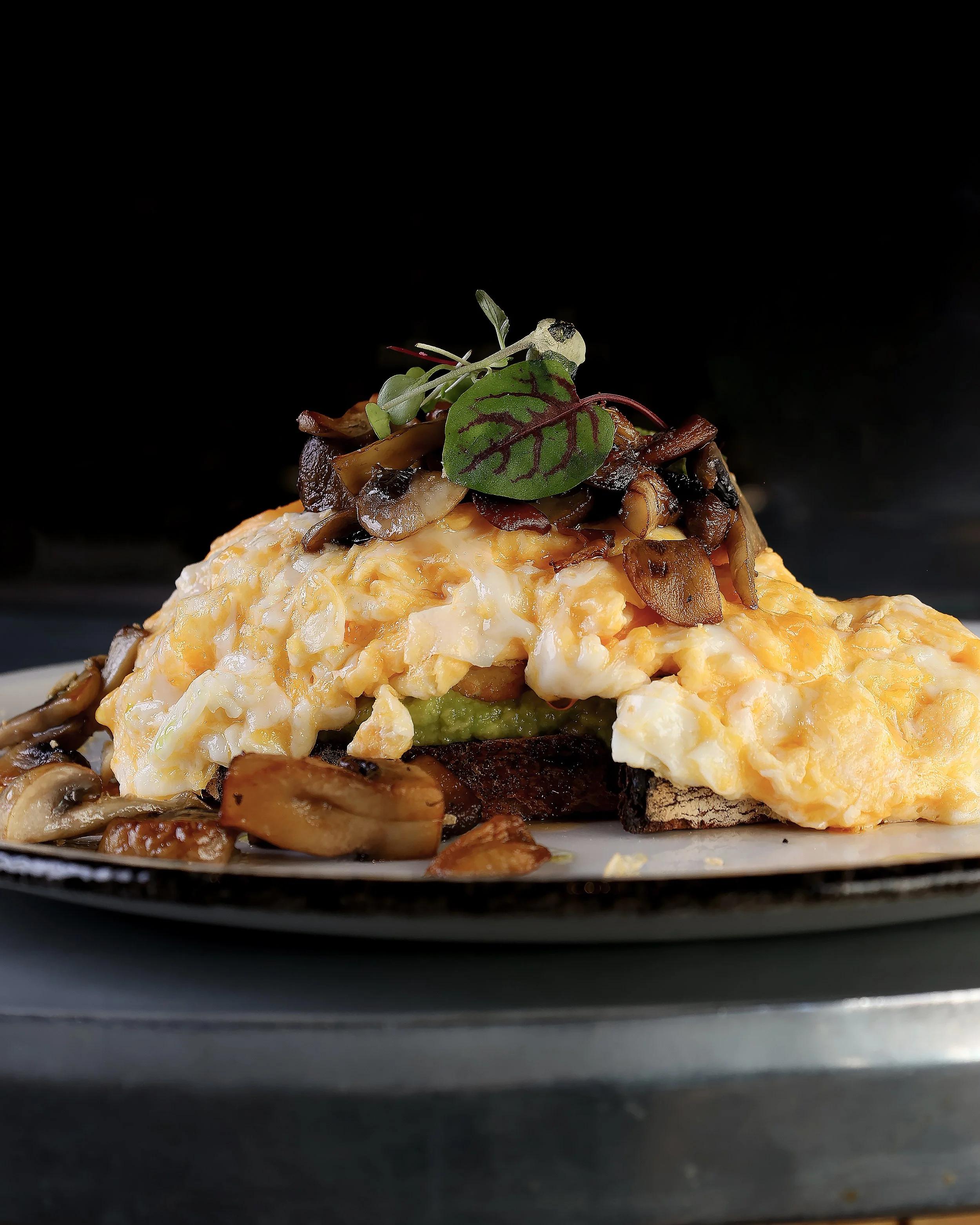 Close-up of a gourmet dish with scrambled eggs, sautéed mushrooms, and microgreens on top, on a black background.