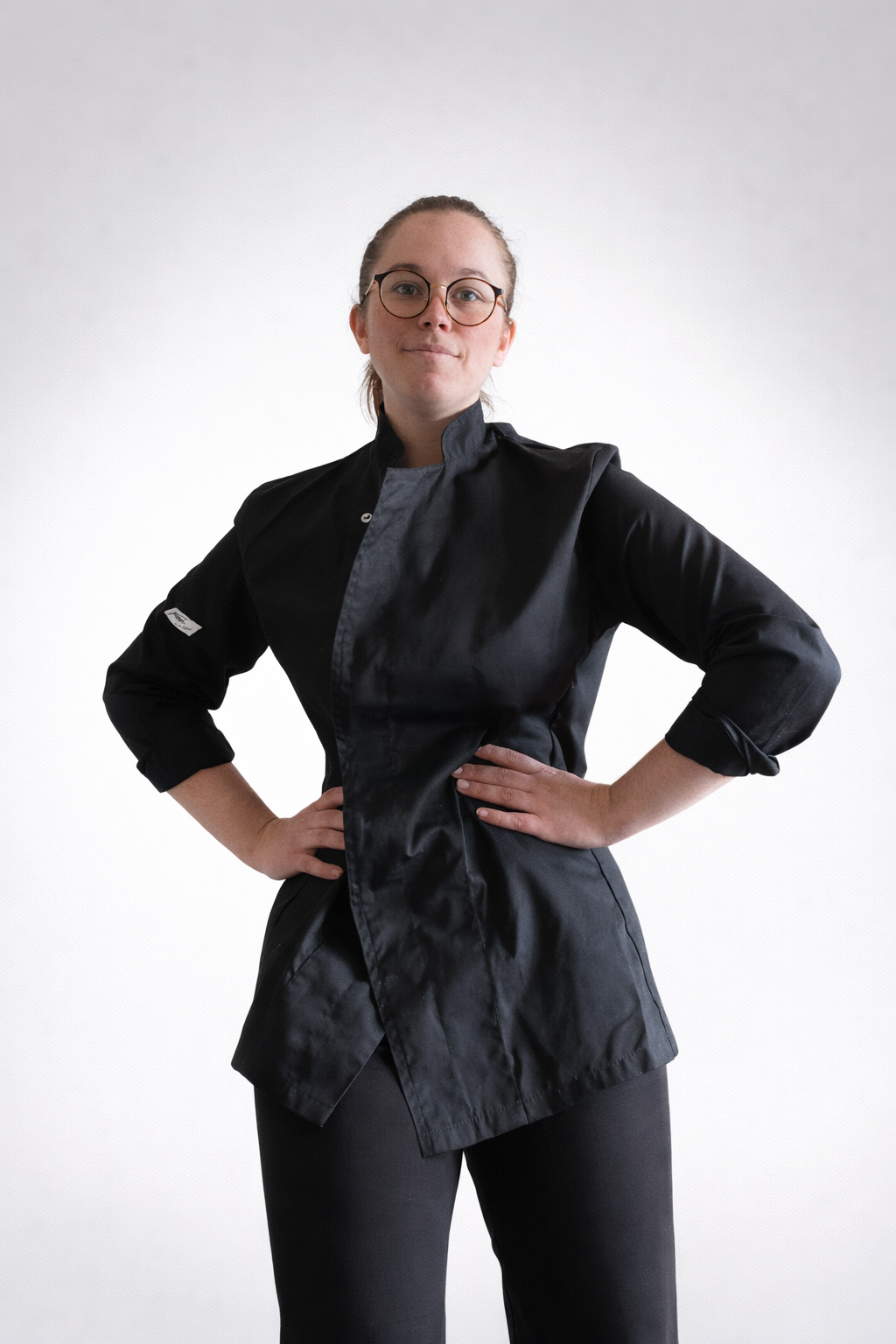 Une femme en uniforme de chef cuisinier se tient avec les mains sur les hanches, souriante, contre un fond blanc.