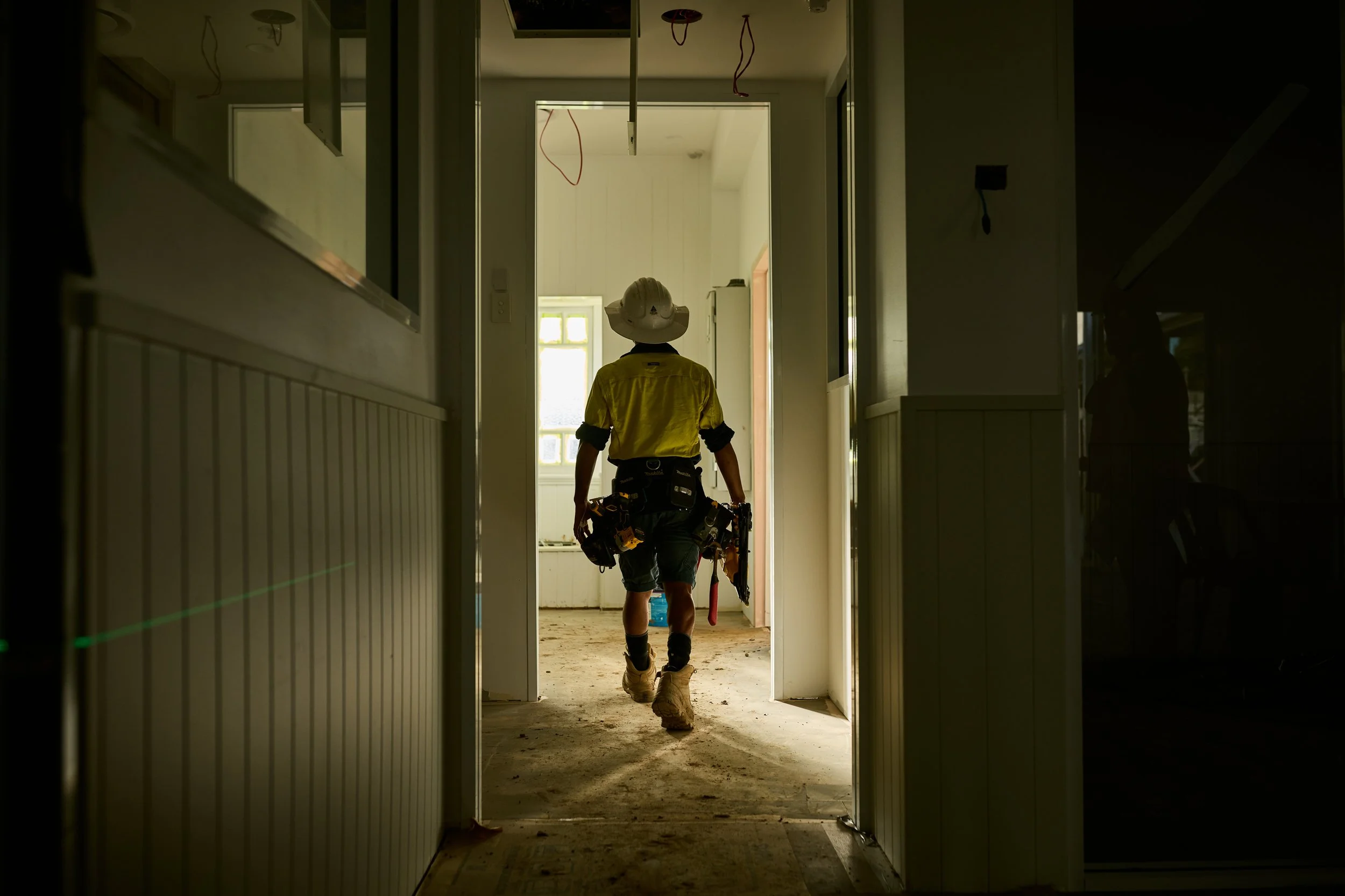 A construction worker wearing a yellow shirt, helmet, and tool belt walking through a doorway in an under-construction building with exposed wires hanging from the ceiling.