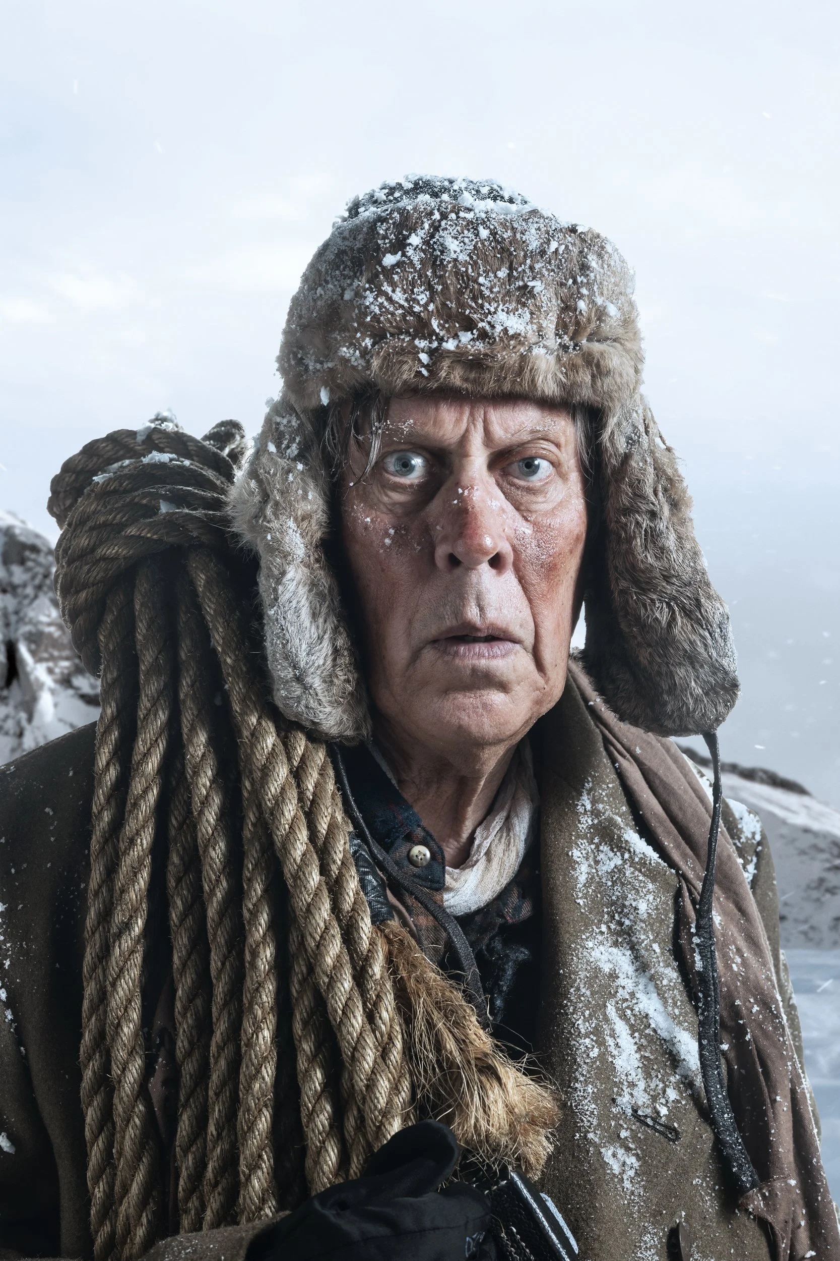 Close-up of a man wearing a heavy, fur-lined hat and a brown jacket in a snowy outdoor setting.